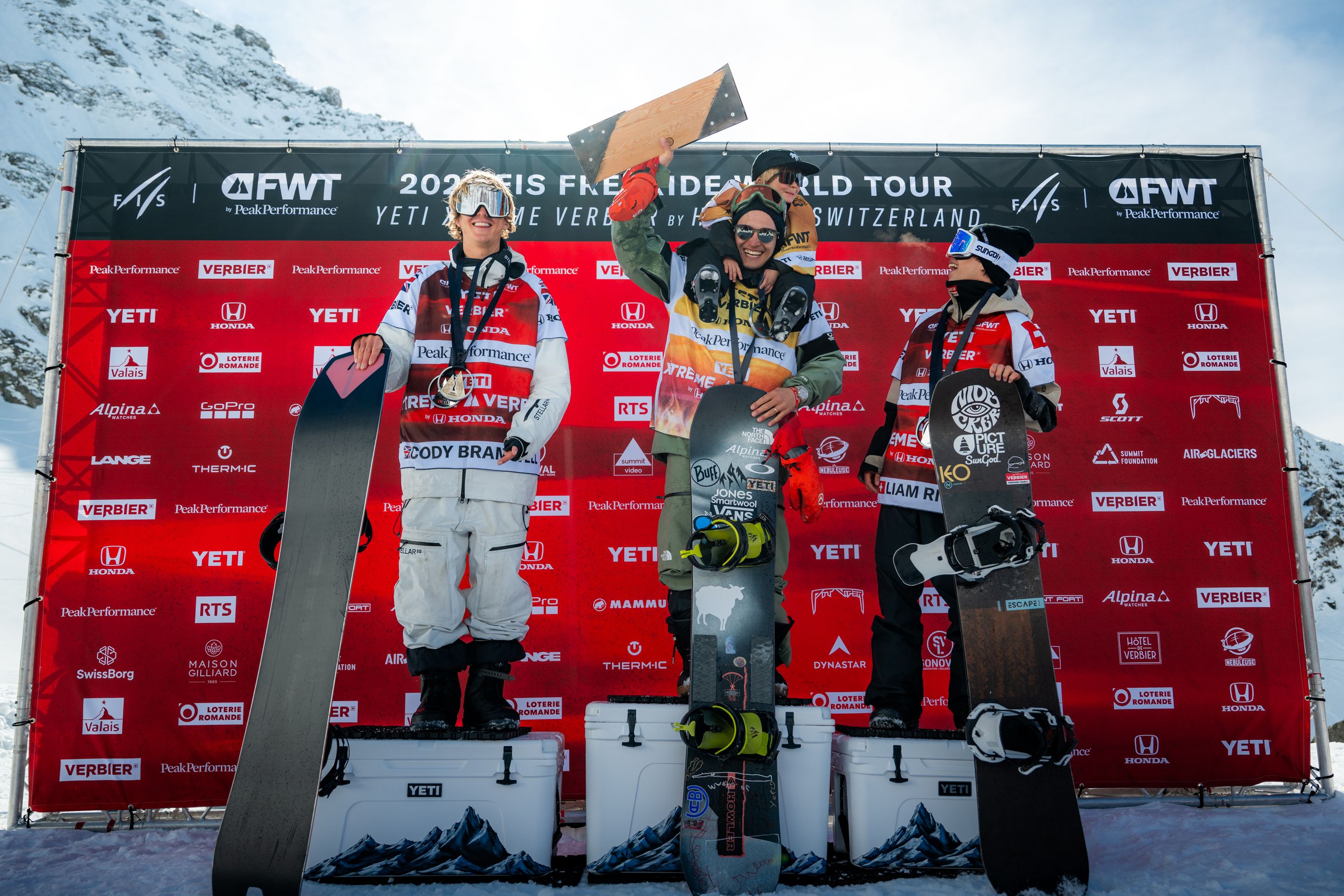Three snowboarders stand on a podium holding snowboards and trophies, celebrating at the FIS Freeride World Tour event in Verbier, Switzerland.