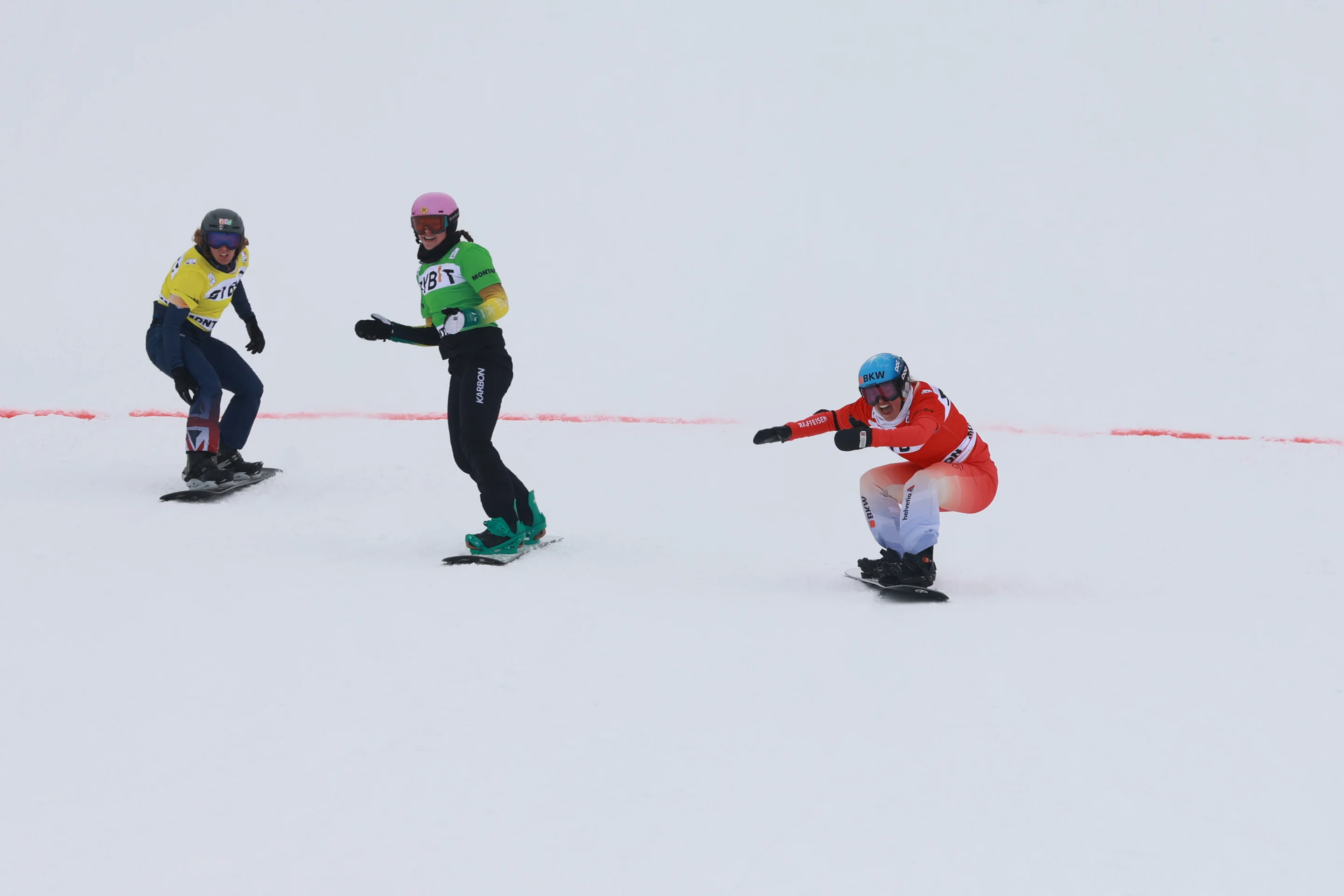 Sina Siegenthaler, in a red bib and snowsuit, crosses the line in front of Josie Baff in a green bib and Charlotte Bankes in a yellow bib. Siegenthaler is crouched down and cheering, Baff and Bankes are upright on their boards.
