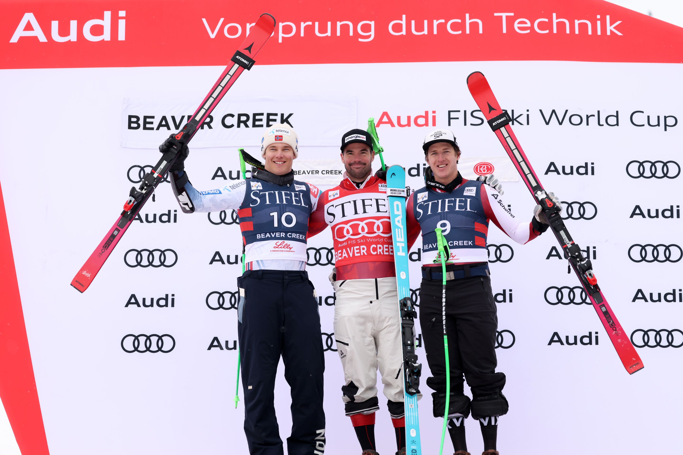 Three skiers on a podium holding their skis overhead in front of an Audi and Beaver Creek FIS Ski World Cup backdrop.