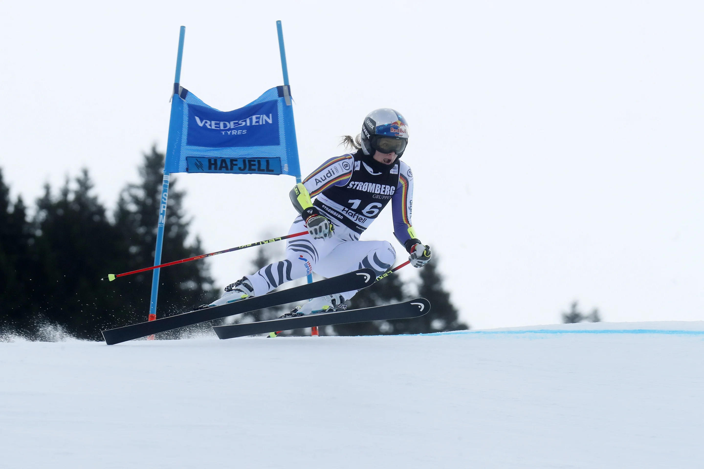 A skier in a black and white uniform and helmet races downhill, navigating around a blue gate with trees in the background.