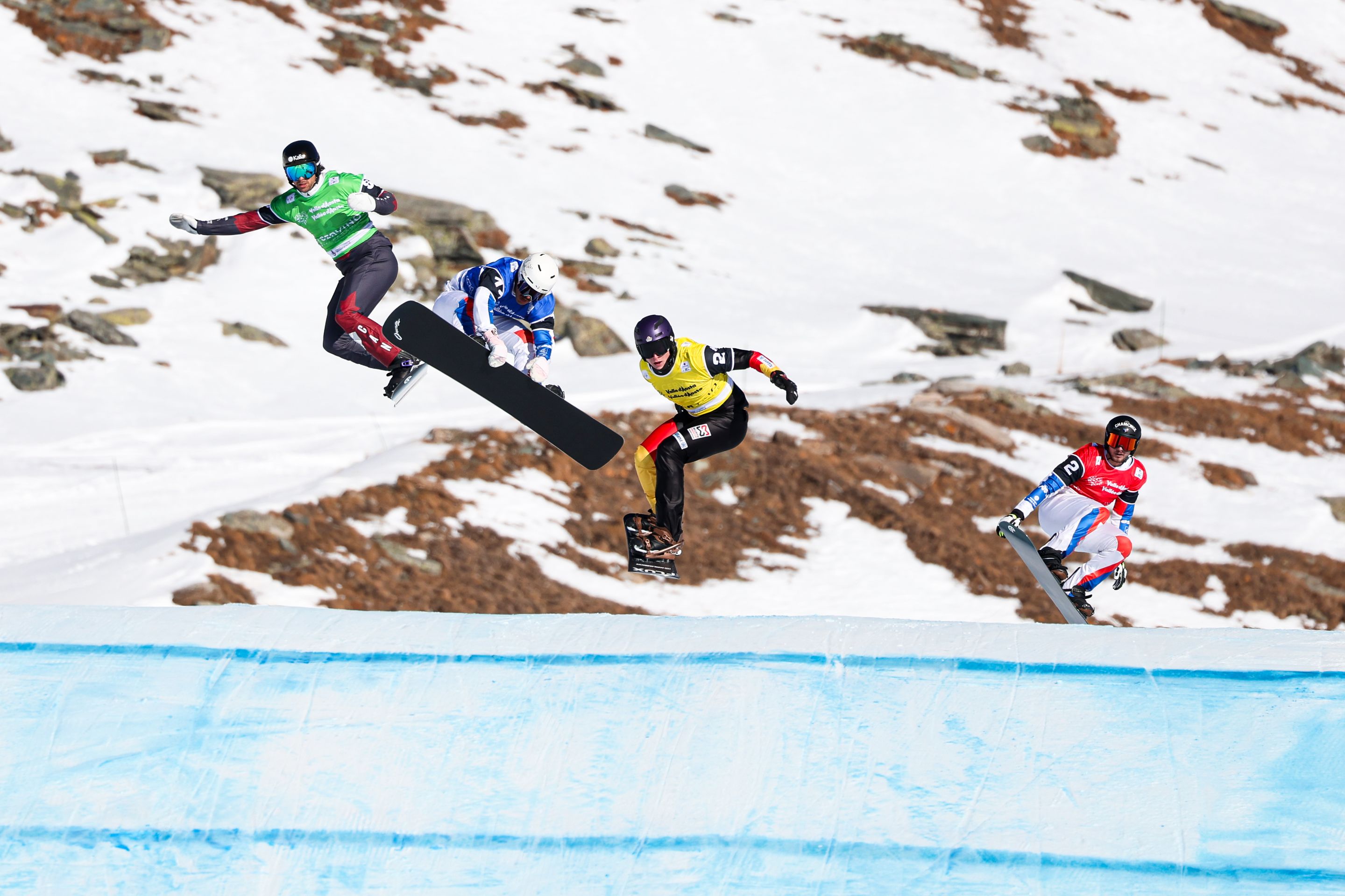 Four snowboarders in coloured bibs race against a mountain backdrop, with a rider in a blue bib grabbing his board