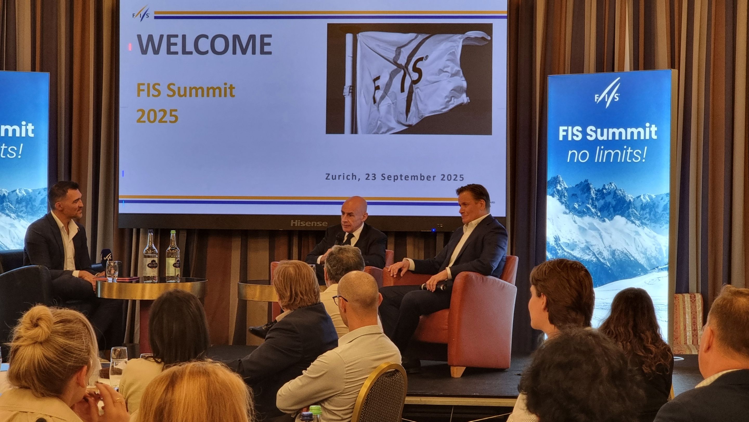 Three men in suits sit on stage discussing at the FIS Summit 2025 in Zurich. A screen displays the event name and date. Audience listens attentively.