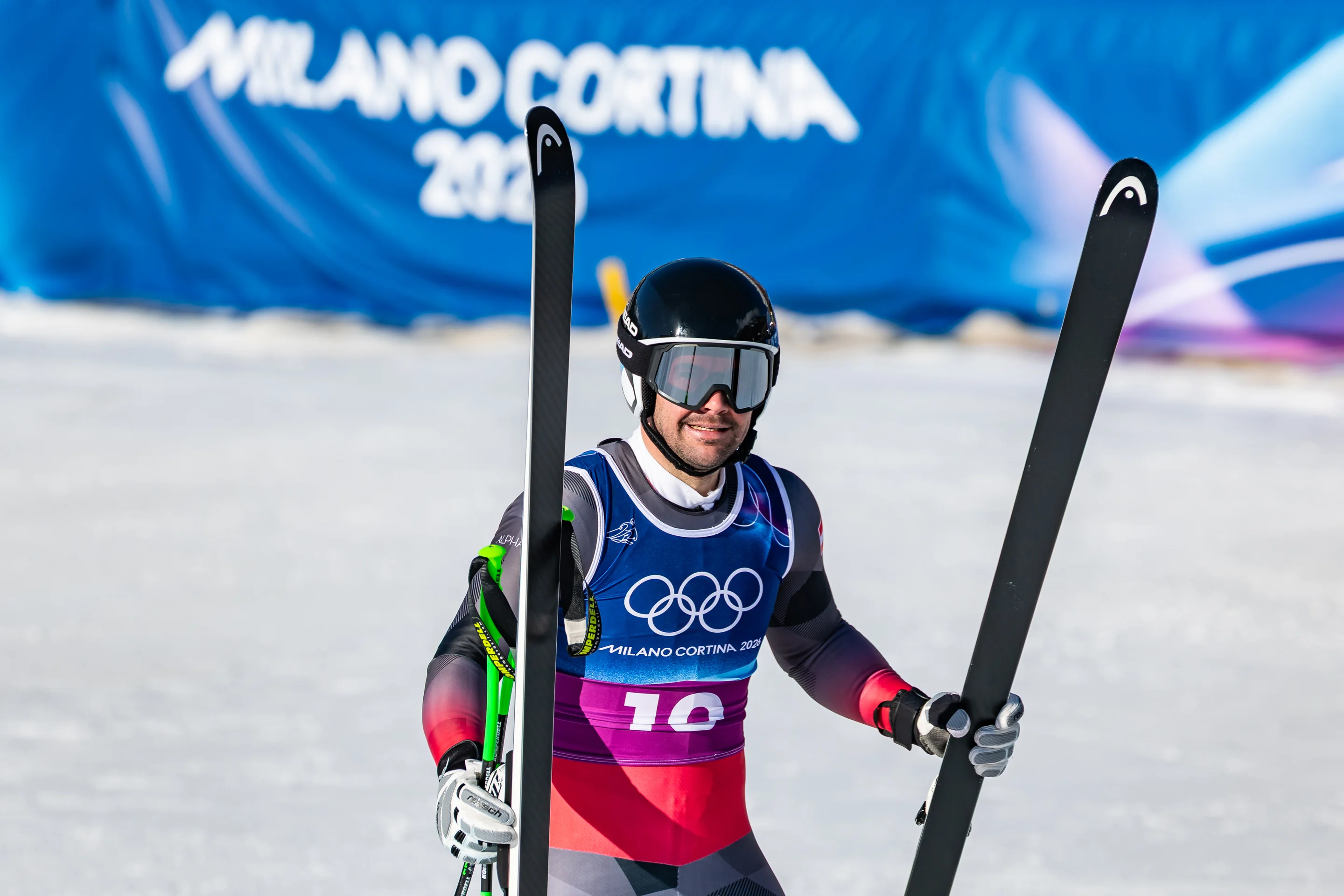 Skier in racing gear holds skis, smiling on a snowy slope. Olympic rings visible on bib; Milano Cortina 2026 banner in the background.