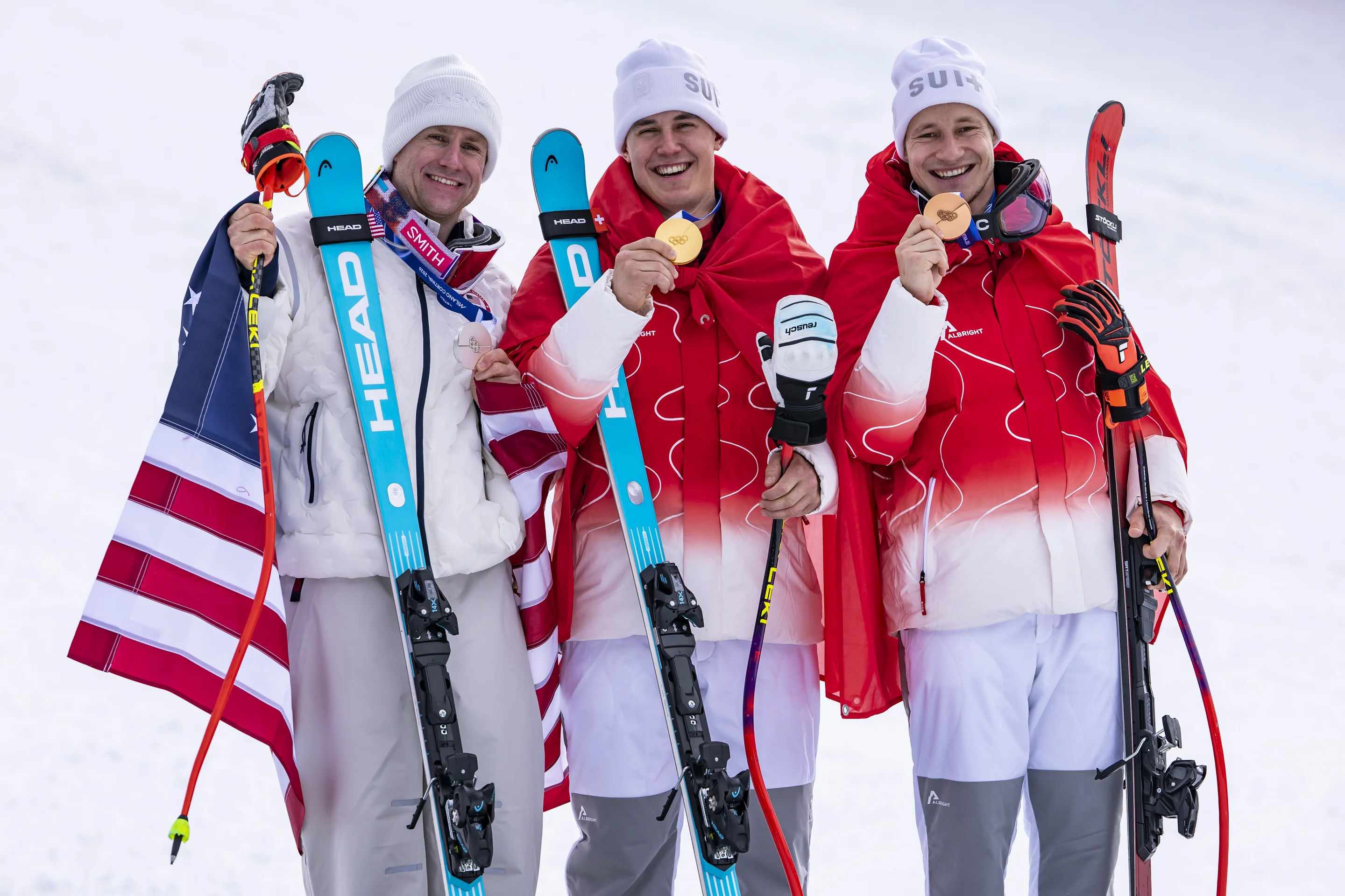 Three skiers in red and white attire hold gold medals and ski poles, smiling on a snowy slope, with one draped in a flag.