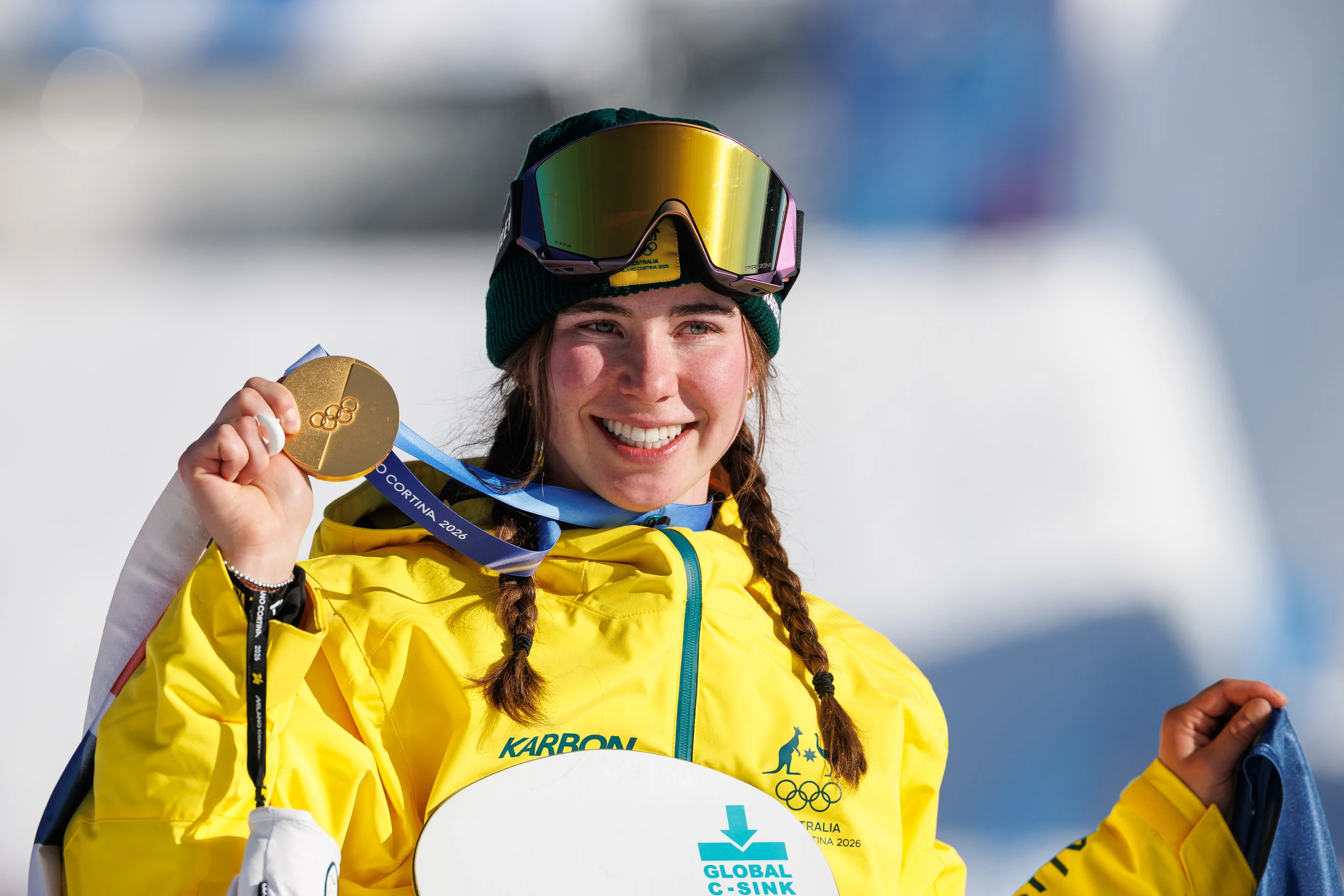 Wearing a yellow tracksuit with ski goggles on her head, Josie Baff of Australia smiles while holding up her Olympic gold medal