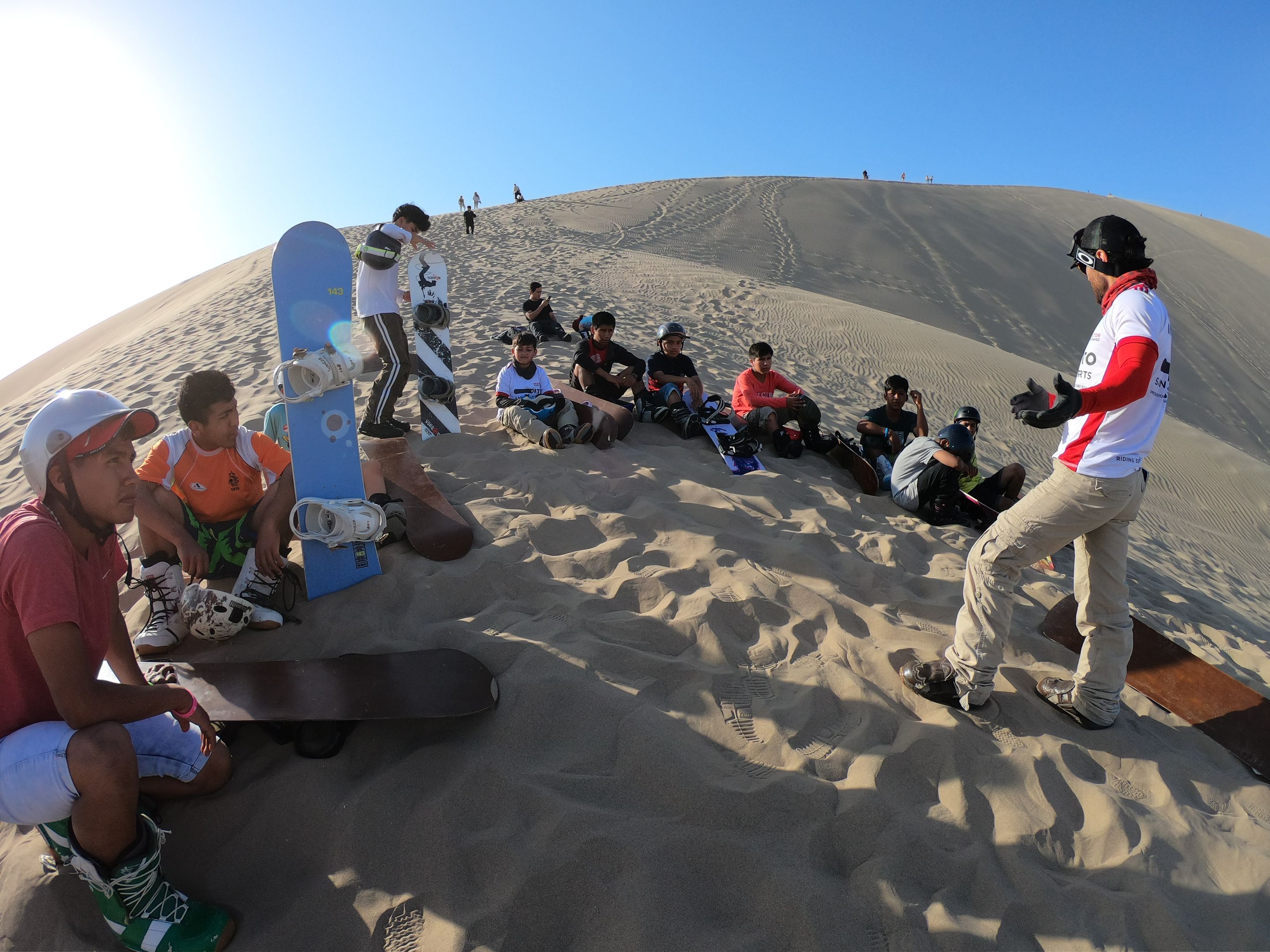 A group of sandboarders sit on top of a dune listening to Dito Chavez, in a white and red t-shirt, explain the sport. The sky is blue behind them.