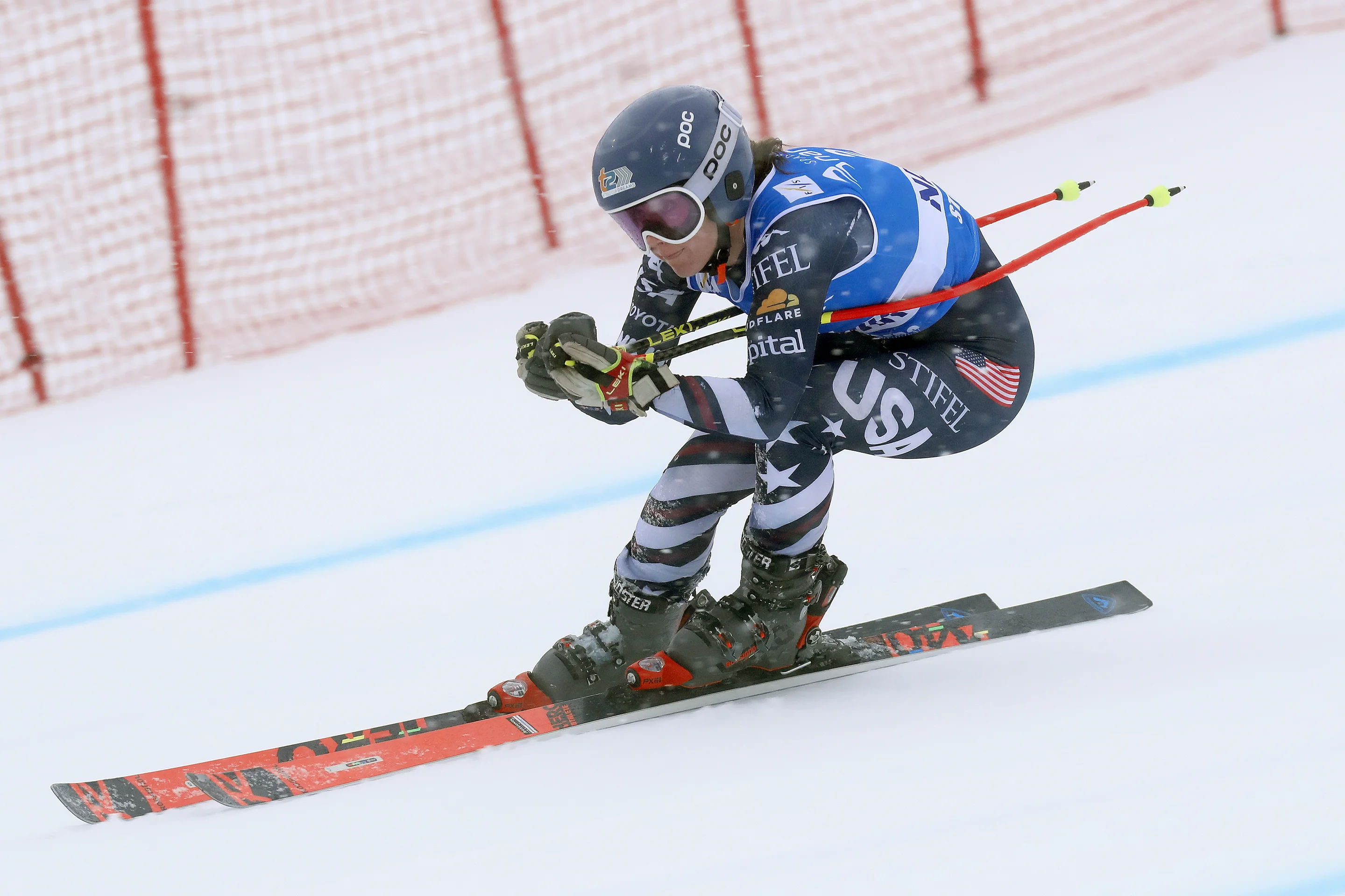 A skier in a blue and black suit races downhill, wearing a helmet and goggles, with orange and black skis, and holding poles in a crouched position.