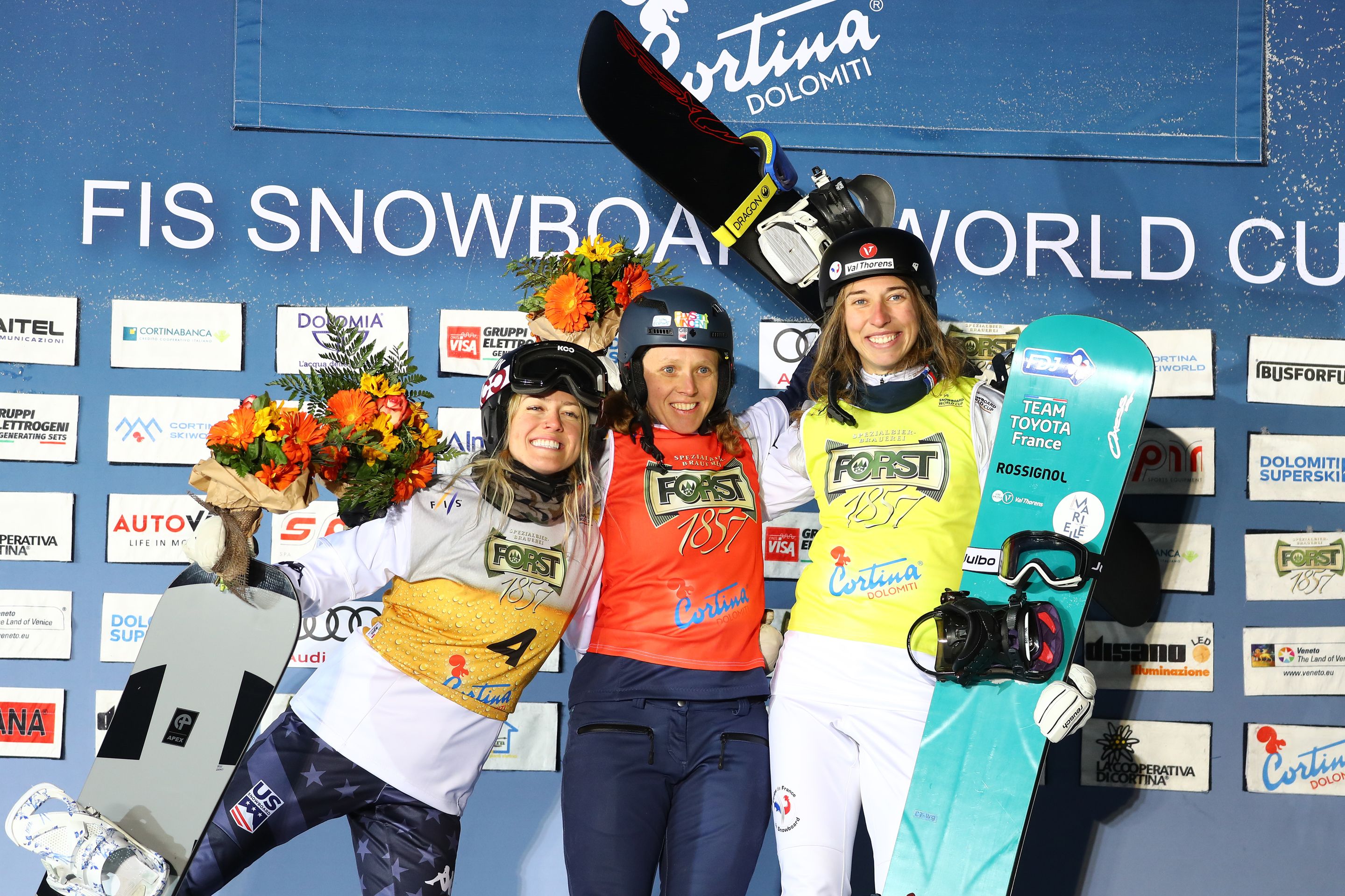 Faye Thelen, in a bib that looks like a glass of beer, celebrates second place at the World Cup in Cortina in February 2023, alongside Charlotte Bankes in a red bib and Chloe Trespeuch in yellow
