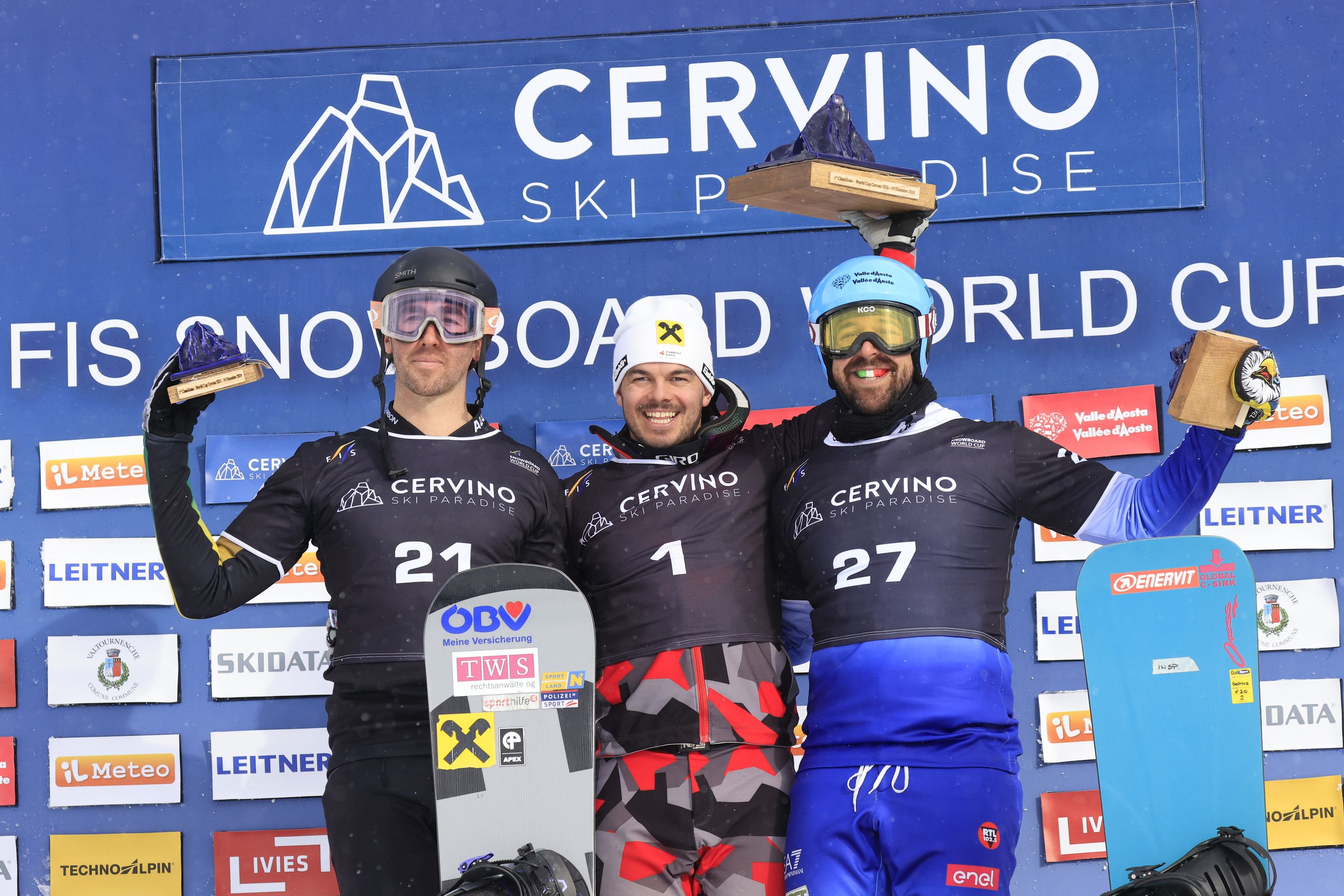 Three snowboarders on a podium raise their trophies in the air, against a backdrop labelled 'Cervino Ski Paradise' with sponsor logos