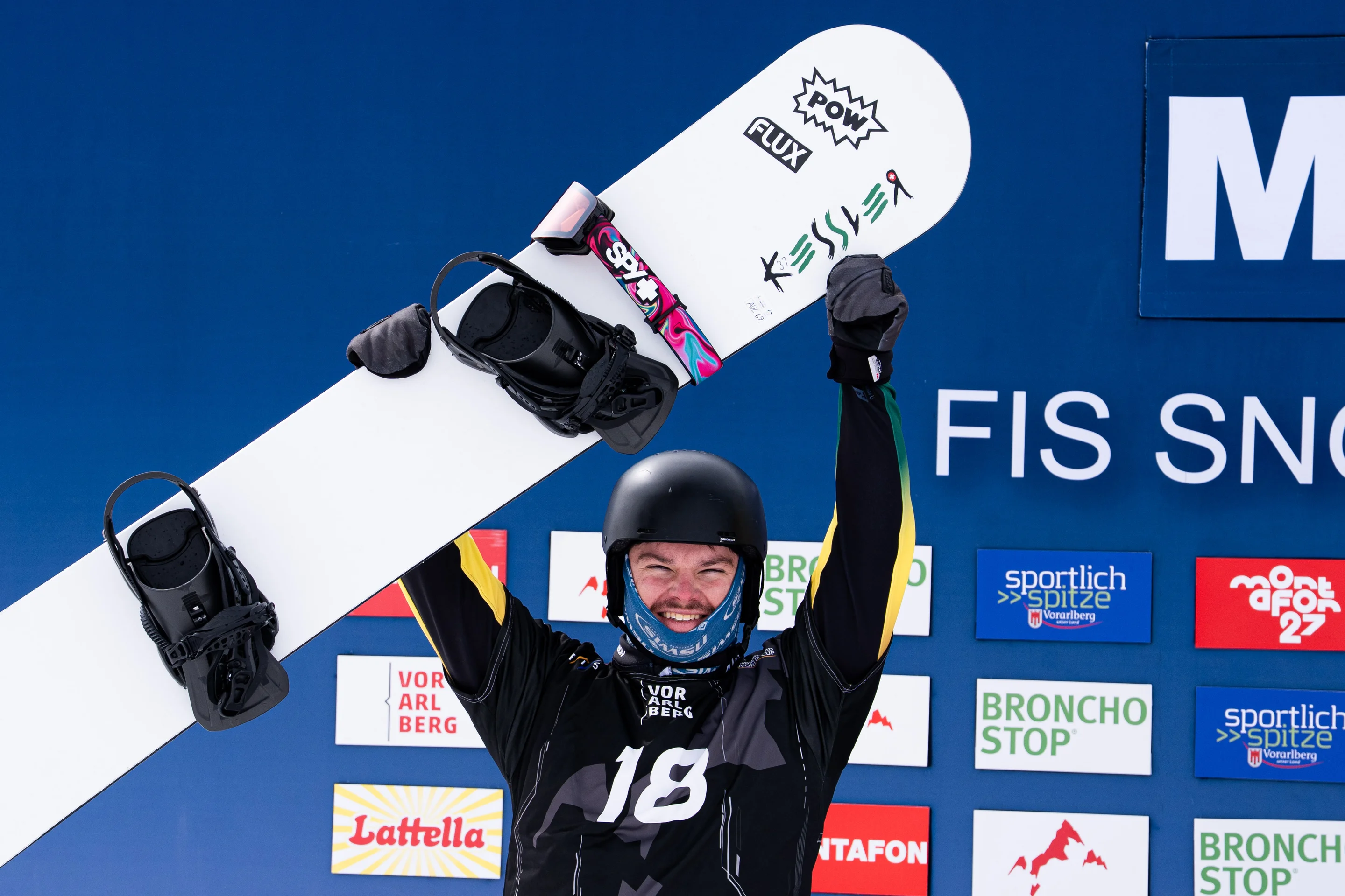 Australian snowboarder Adam Lambert holds his snowboard in the air in celebration, wearing a helmet and a protective buff around his face, in front of a blue backdrop