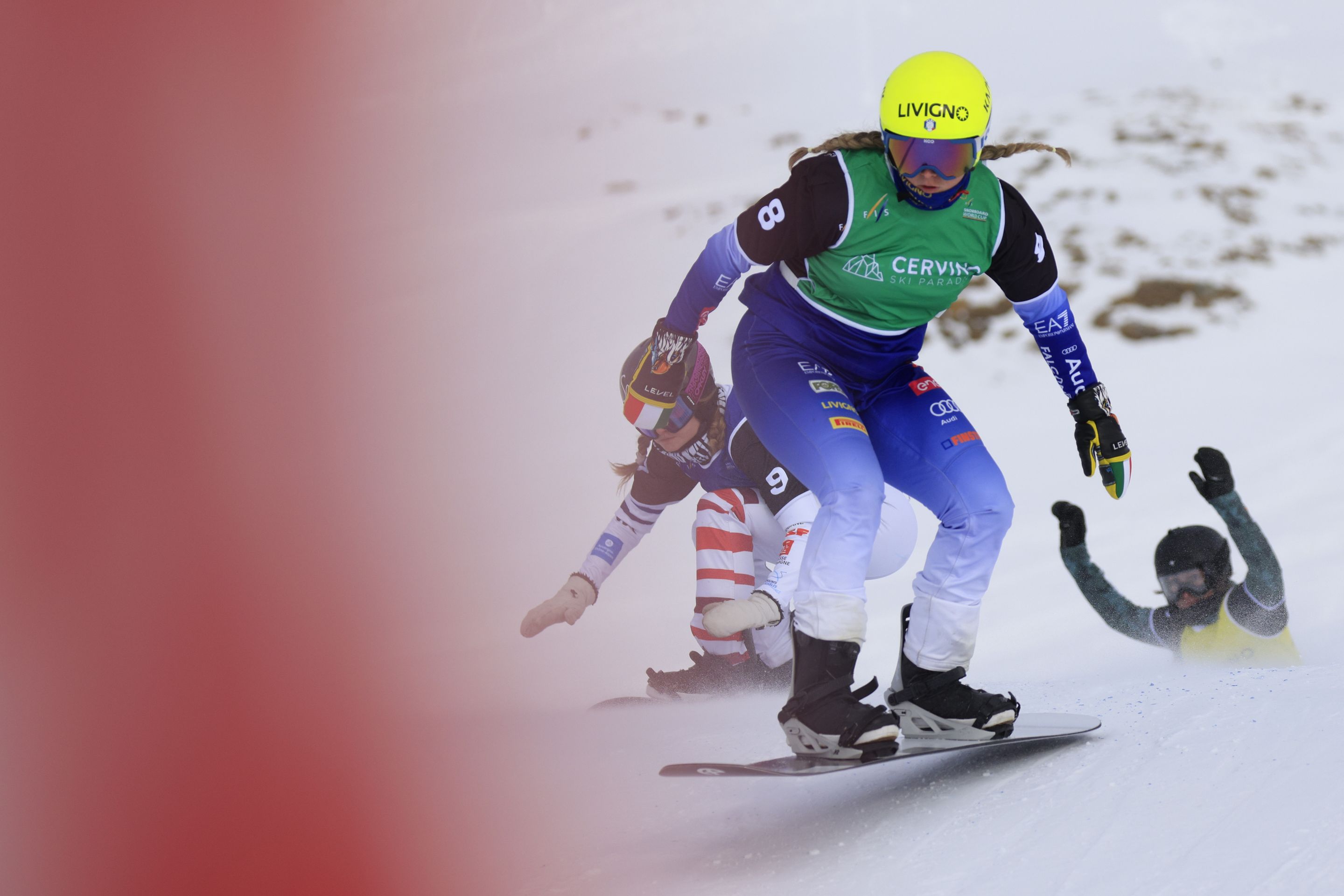 A snowboarder in a green bib and yellow helmet leads a race against a snowy background, her opponents just behind her. Her pigtails are flying in the breeze.