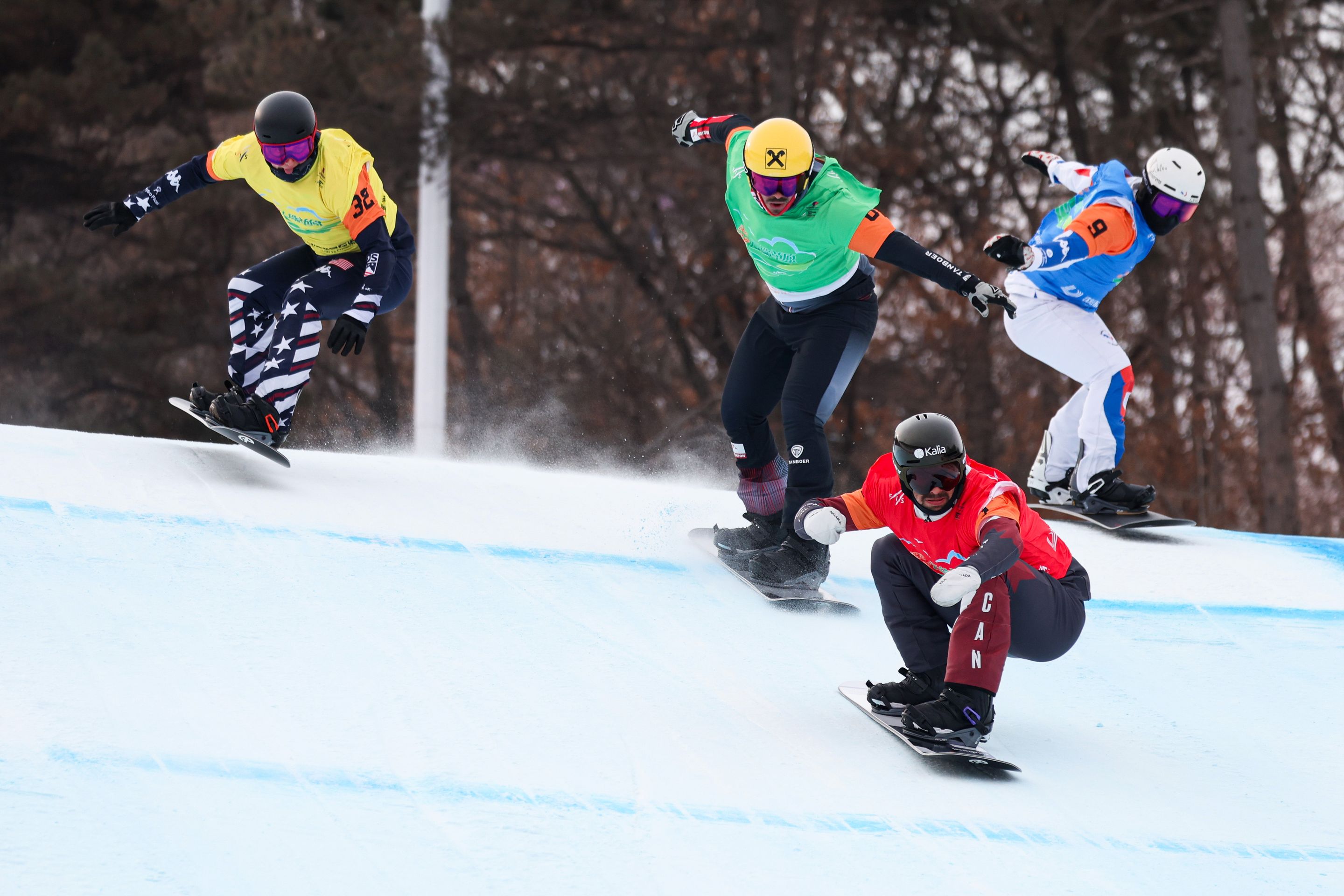 Four snowboarders in colorful gear race down a snowy slope, with trees in the background, during a competitive event.