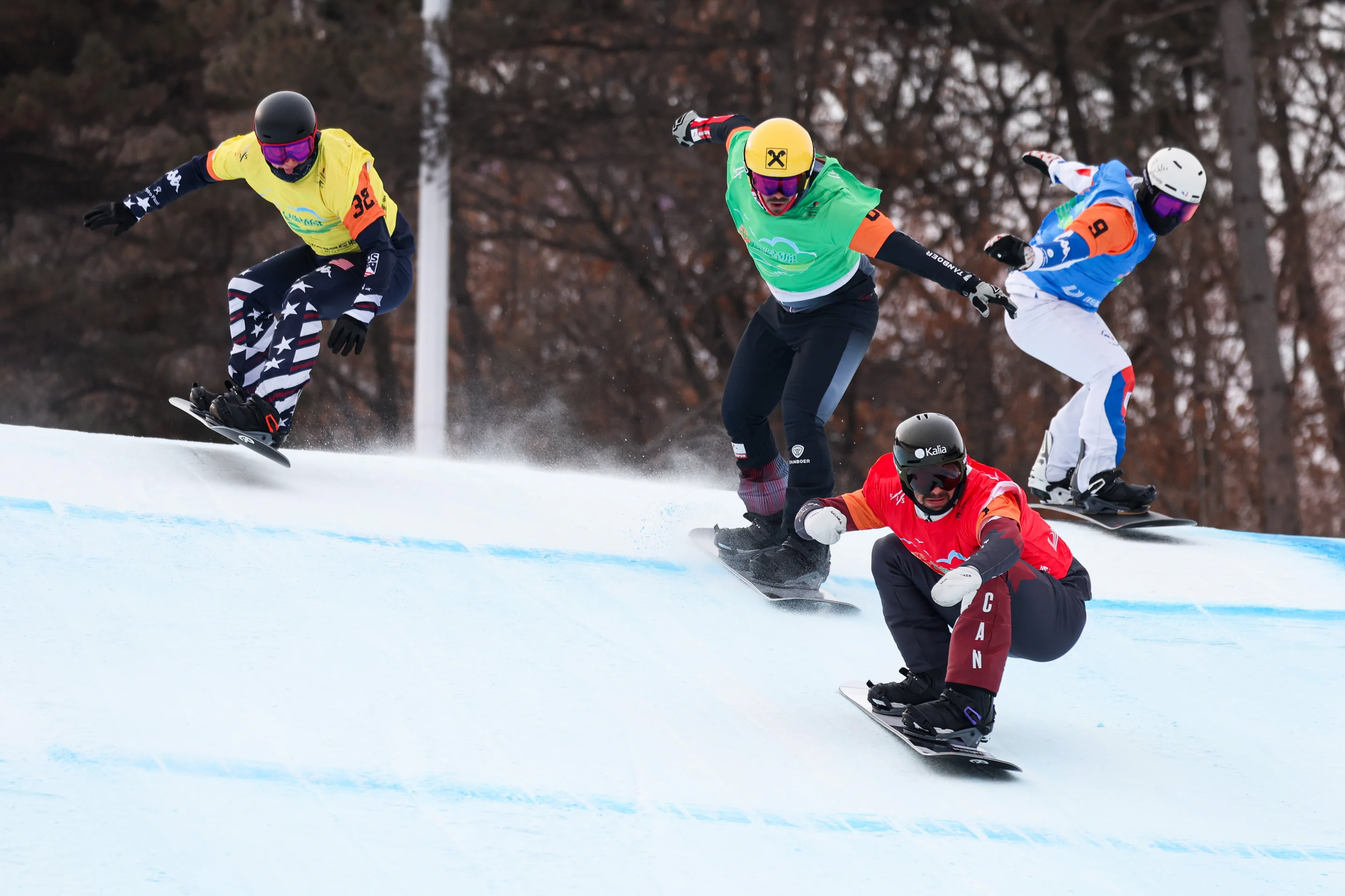Four snowboarders in colorful gear race down a snowy slope, with trees in the background, during a competitive event.