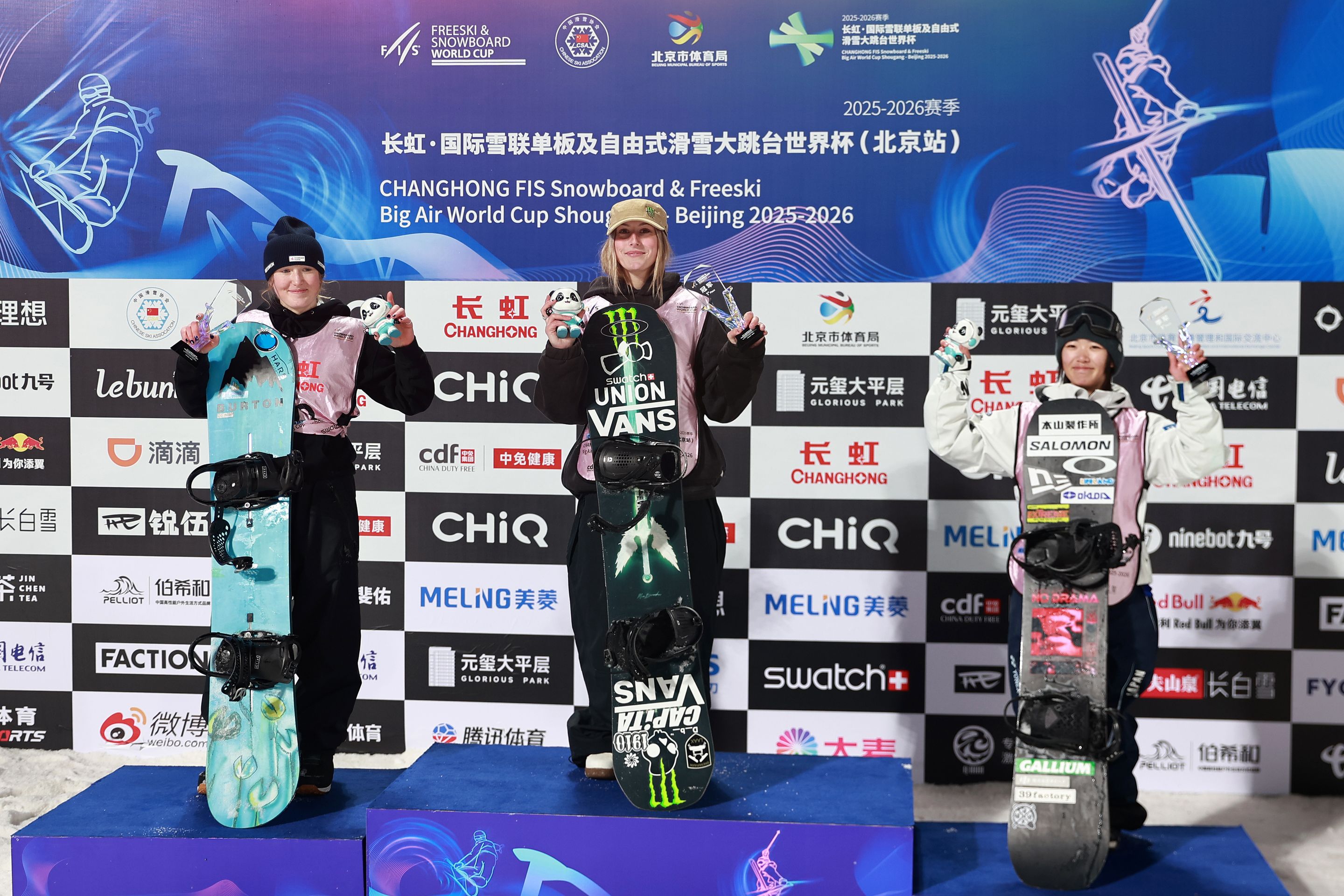 Three snowboarders standing on winners' podium holding snowboards and medals in front of Big Air World Cup sponsor backdrop.