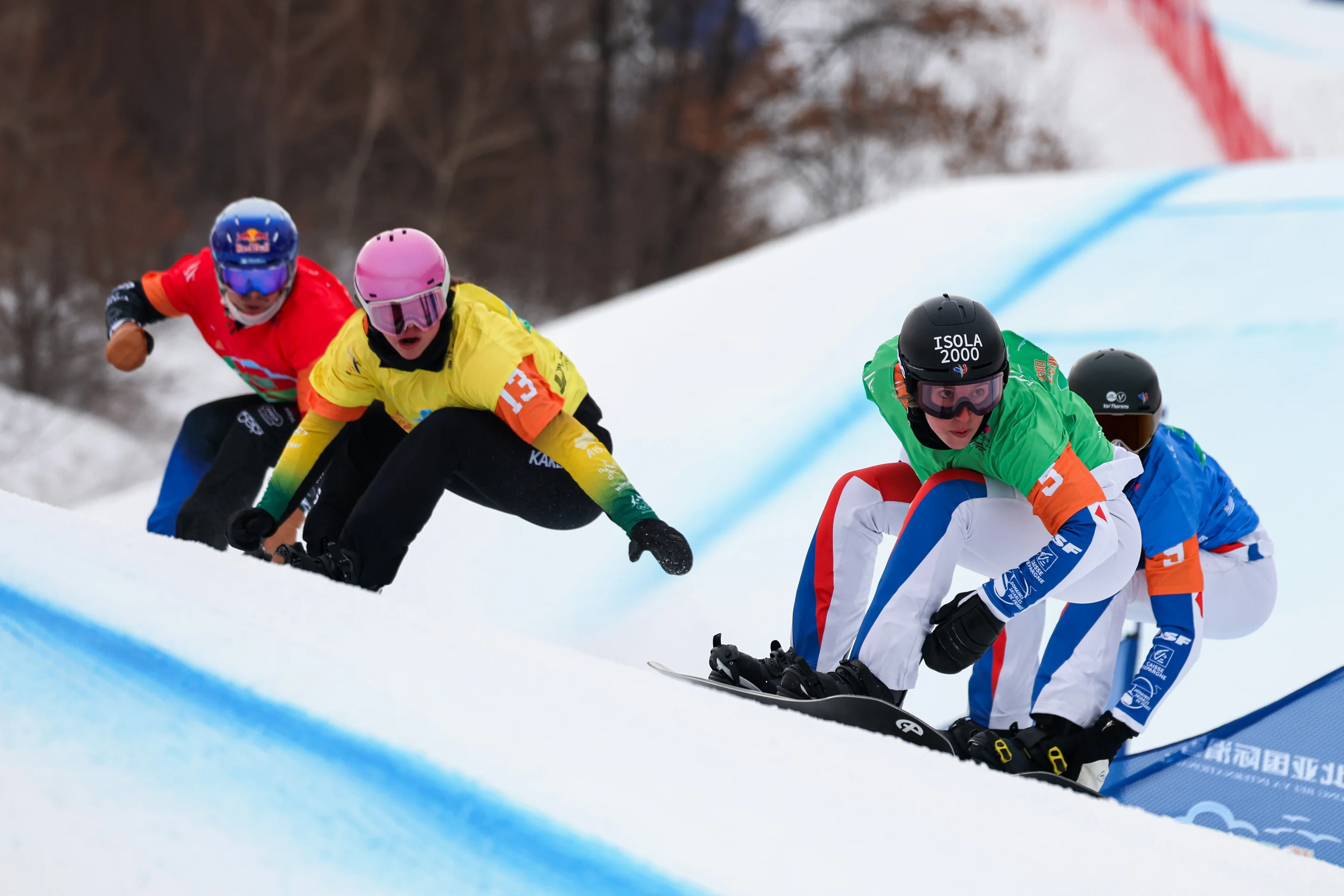 Four snowboarders in colorful gear race down a snowy slope during a snowboarding competition, surrounded by trees.