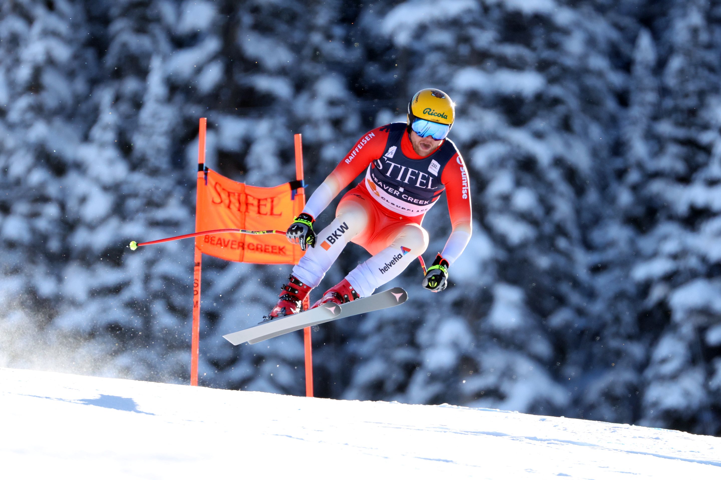 Skier in red racing suit and yellow helmet airborne past an orange gate on a snowy slope with snow-covered pine trees in the background.