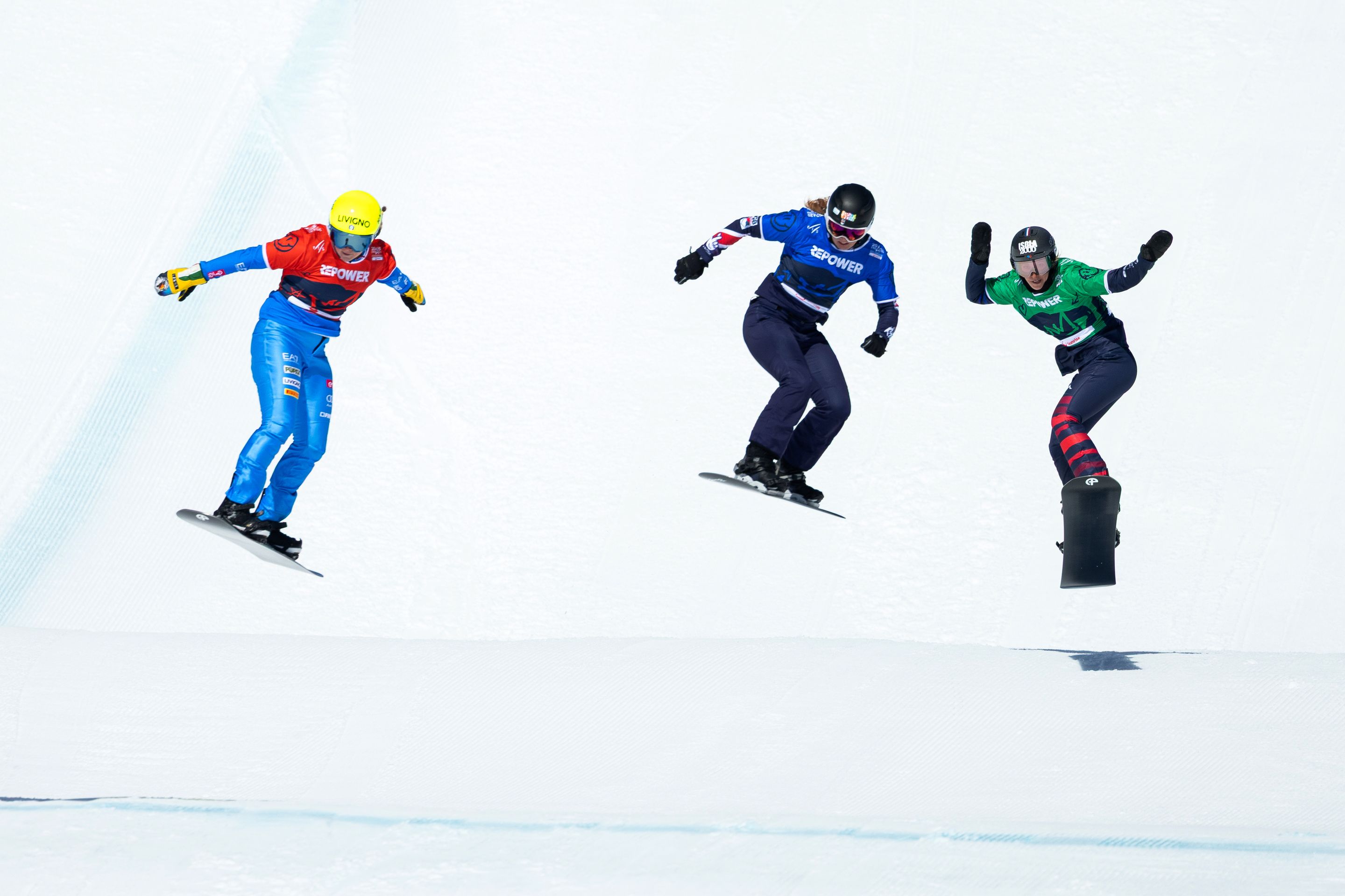 Three snowboarders, in red, blue and green bibs, take a jump at the 2025 World Championships