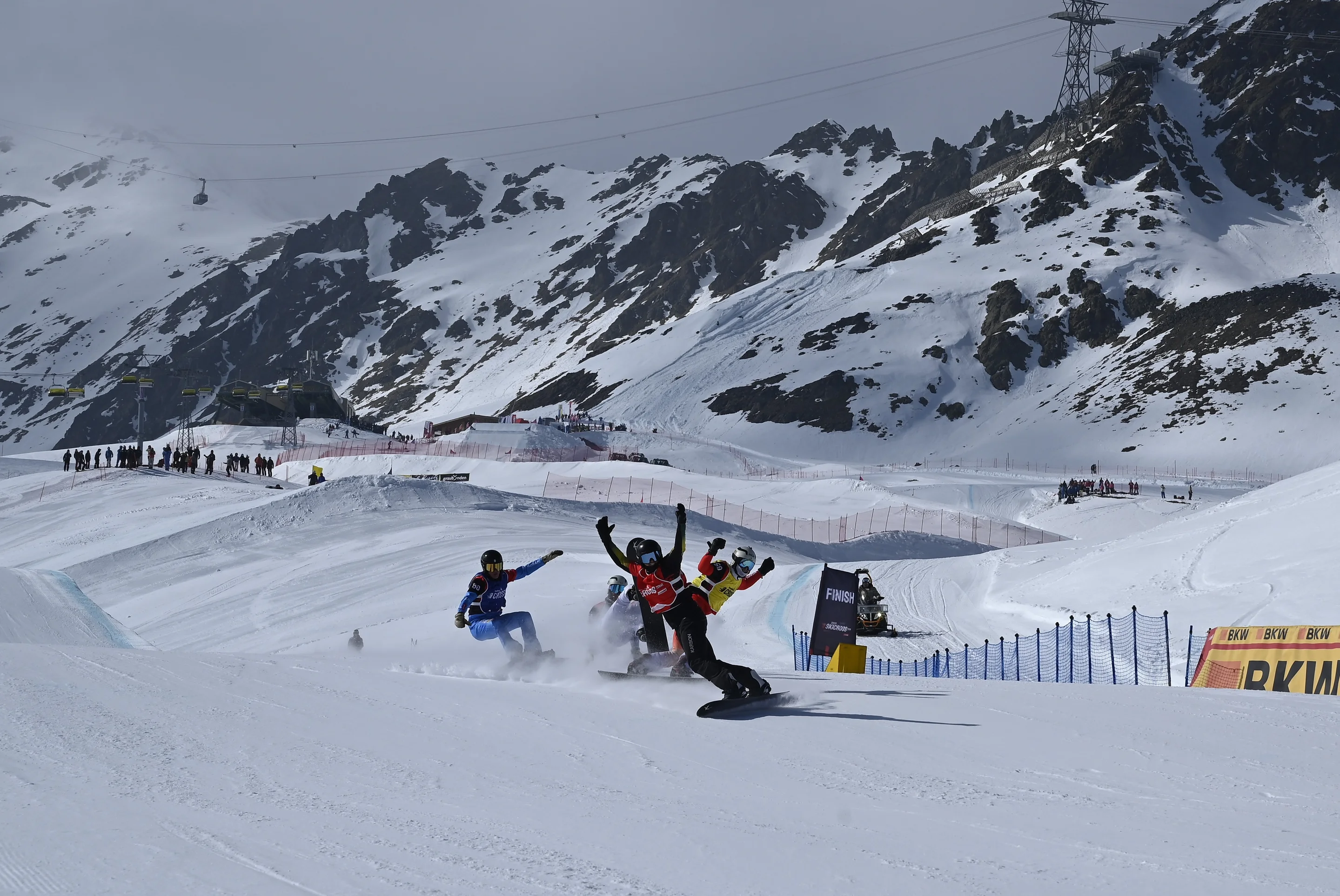 Four snowboarders fight for places as they cross the finish line, with mountains behind them and spectators watching in the background