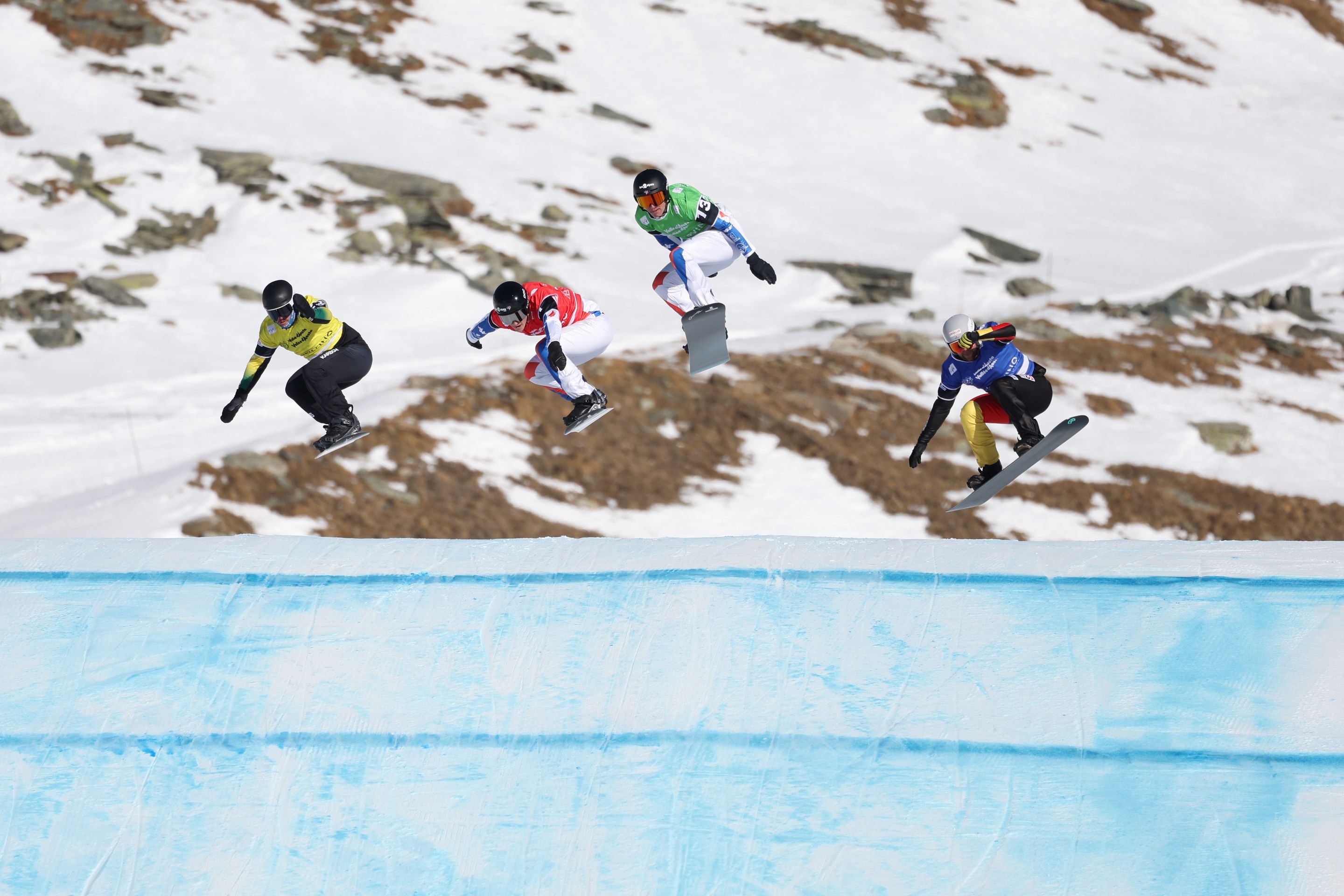 Four snowboarders in coloured bibs take a jump in Cervinia, in front of a snowy mountain background