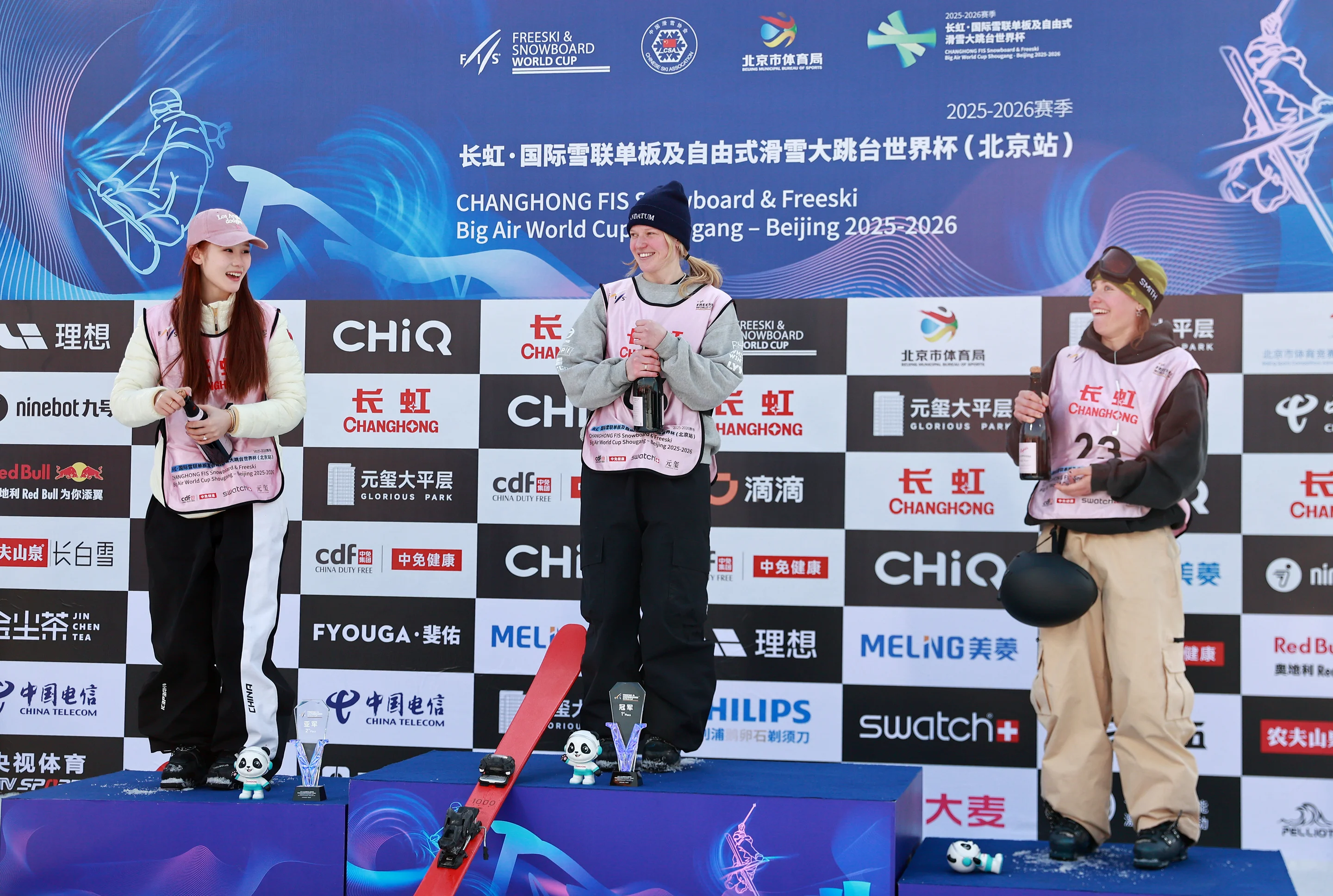 Three female skiers on podium holding trophies at FIS Big Air World Cup in Beijing, sponsor-branded backdrop behind them