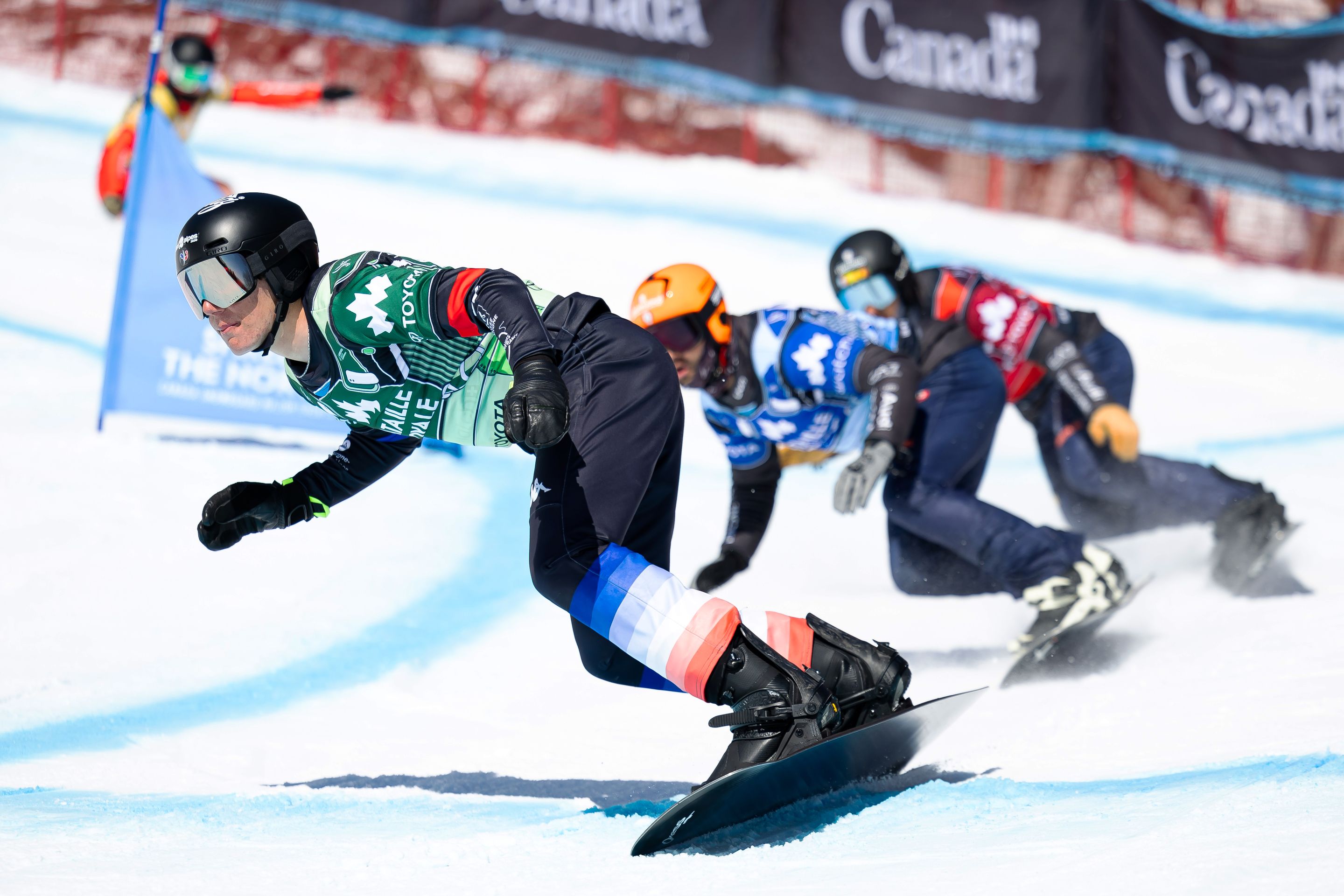 Three snowboarders in a line come around a turn, with Aidan Chollet leading in a green bib