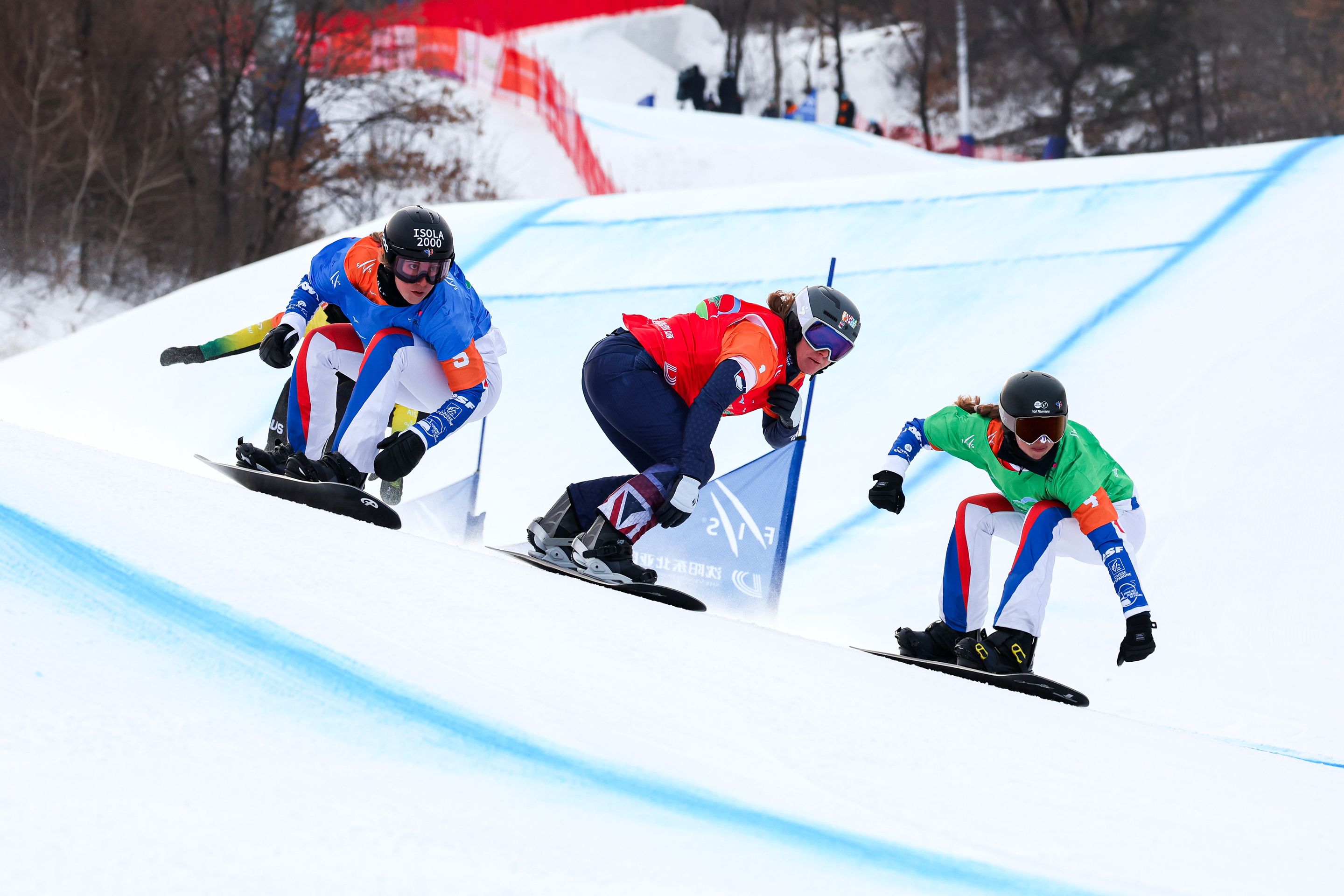 Three snowboarders in colorful gear race down a snowy slope, each focused and leaning into the curve, with trees and a red safety net in the background.