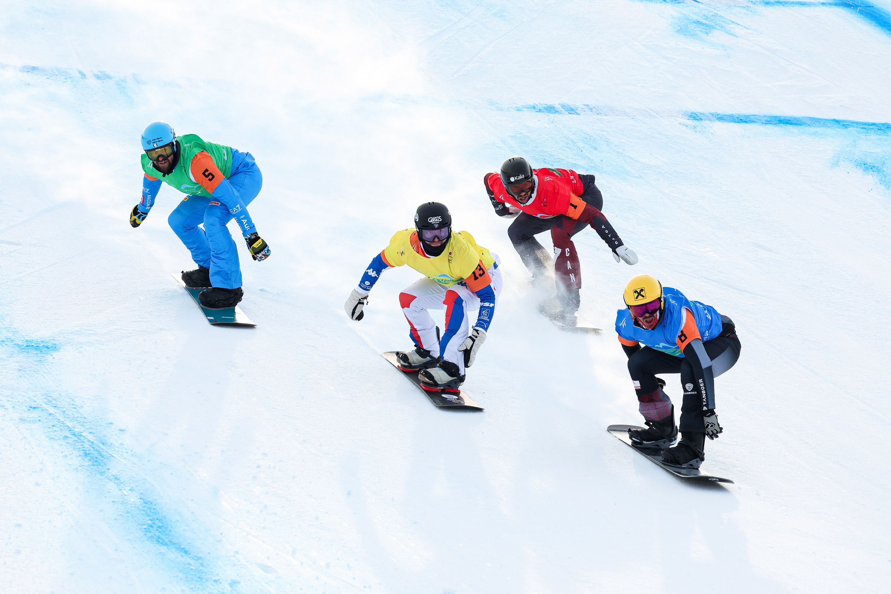 Four snowboarders racing downhill on a snowy course, dressed in colorful gear, with numbered bibs and helmets, on a clear day.