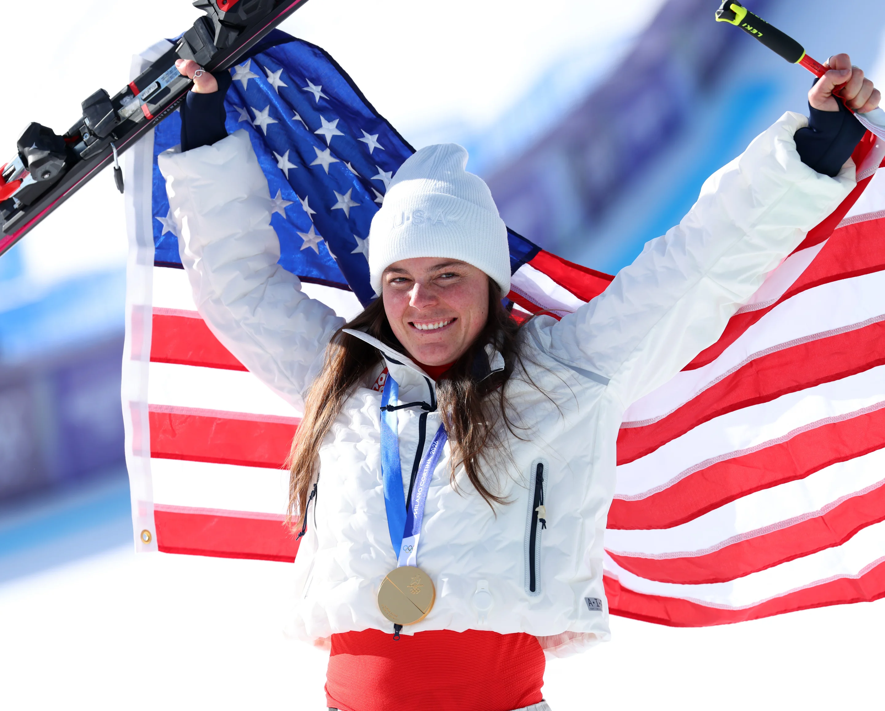 Athlete in a white jacket and beanie celebrates with skis and a large American flag, wearing a medal, against a snowy backdrop.