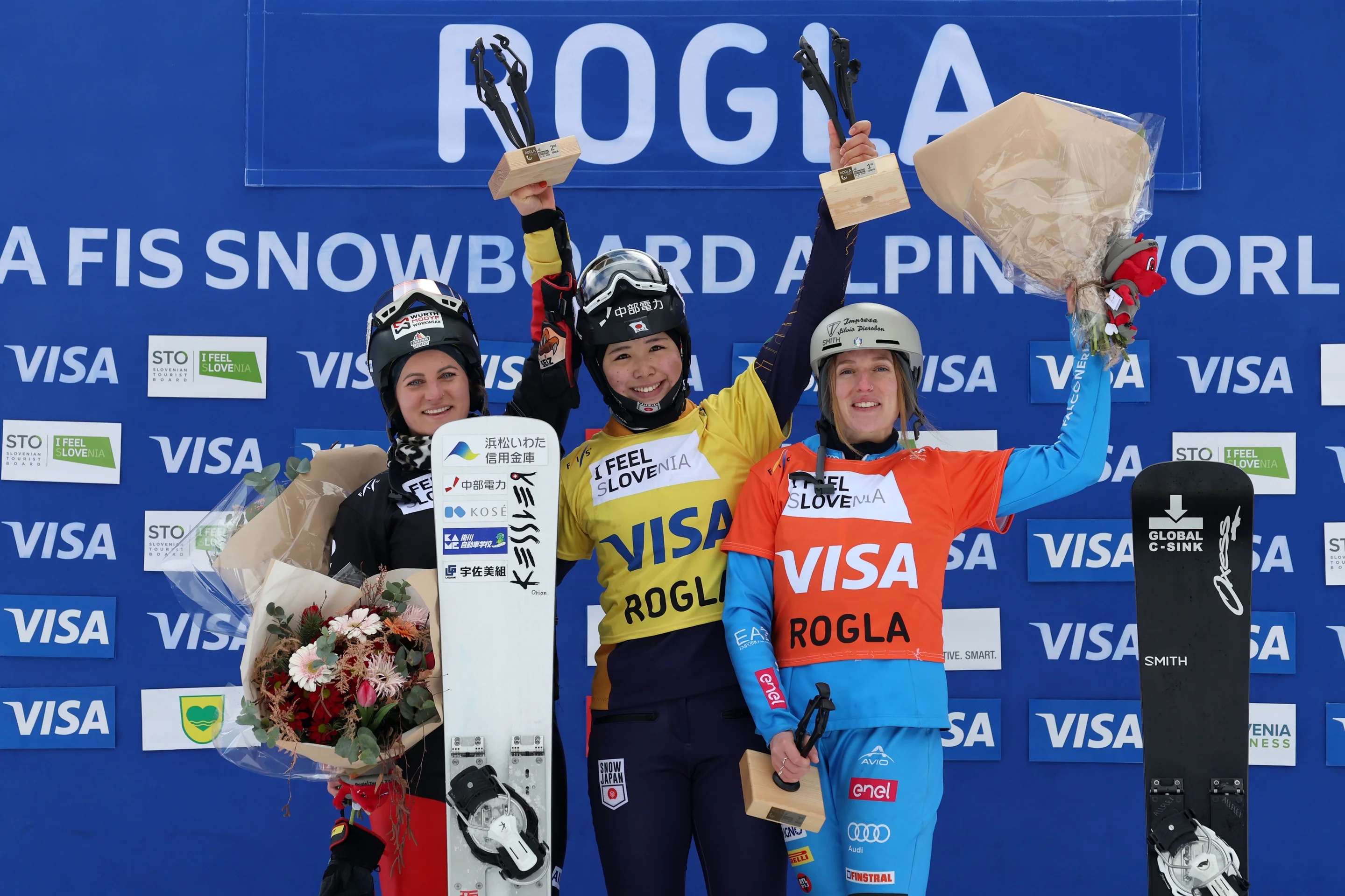 Three snowboarders stand on a podium holding trophies and flowers, celebrating their victory at the FIS Snowboard Alpine World Cup in Rogla.