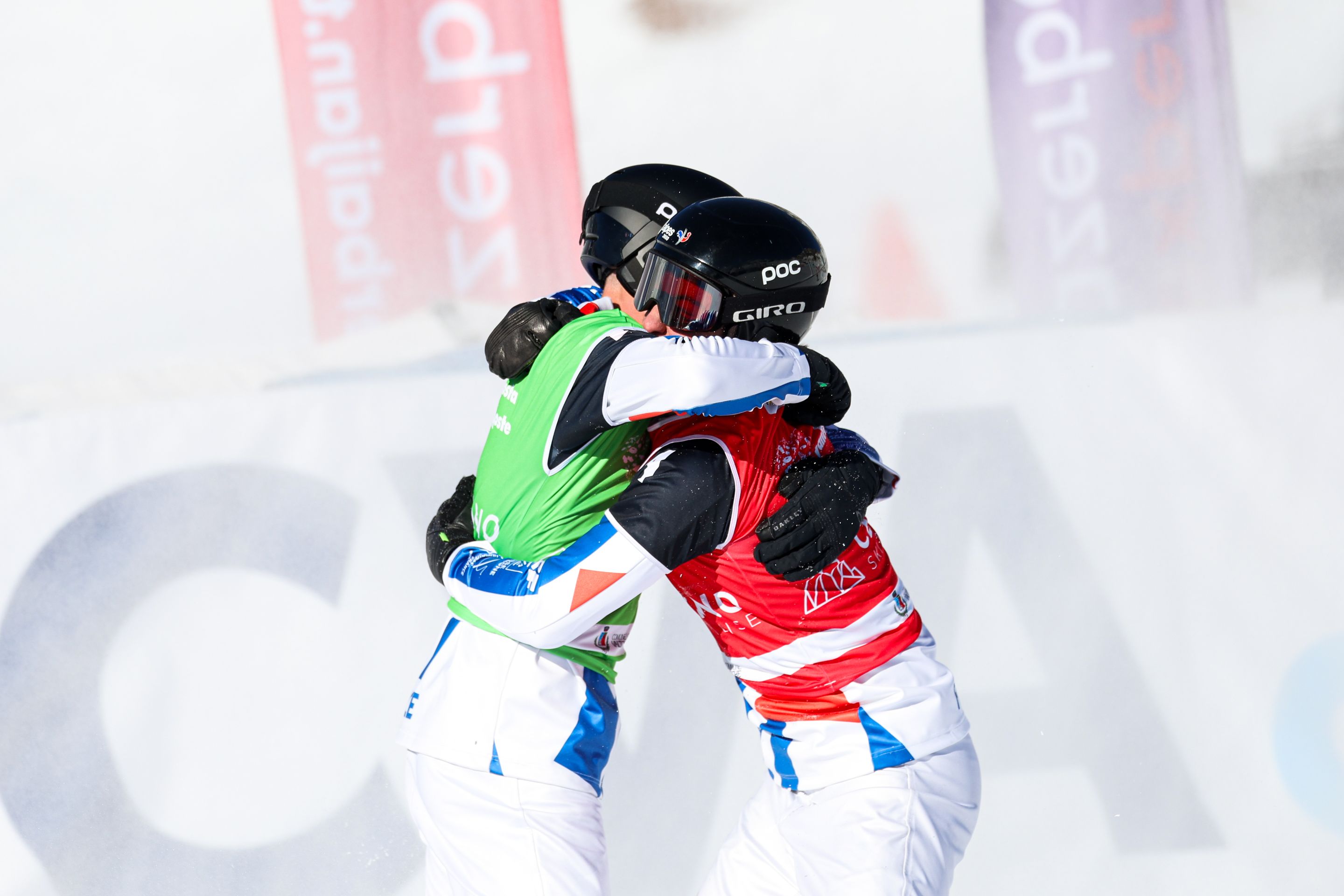 Two snowboarders in green and red bibs and white outfits embrace against a snowy background © FIS/Miha Matavz
