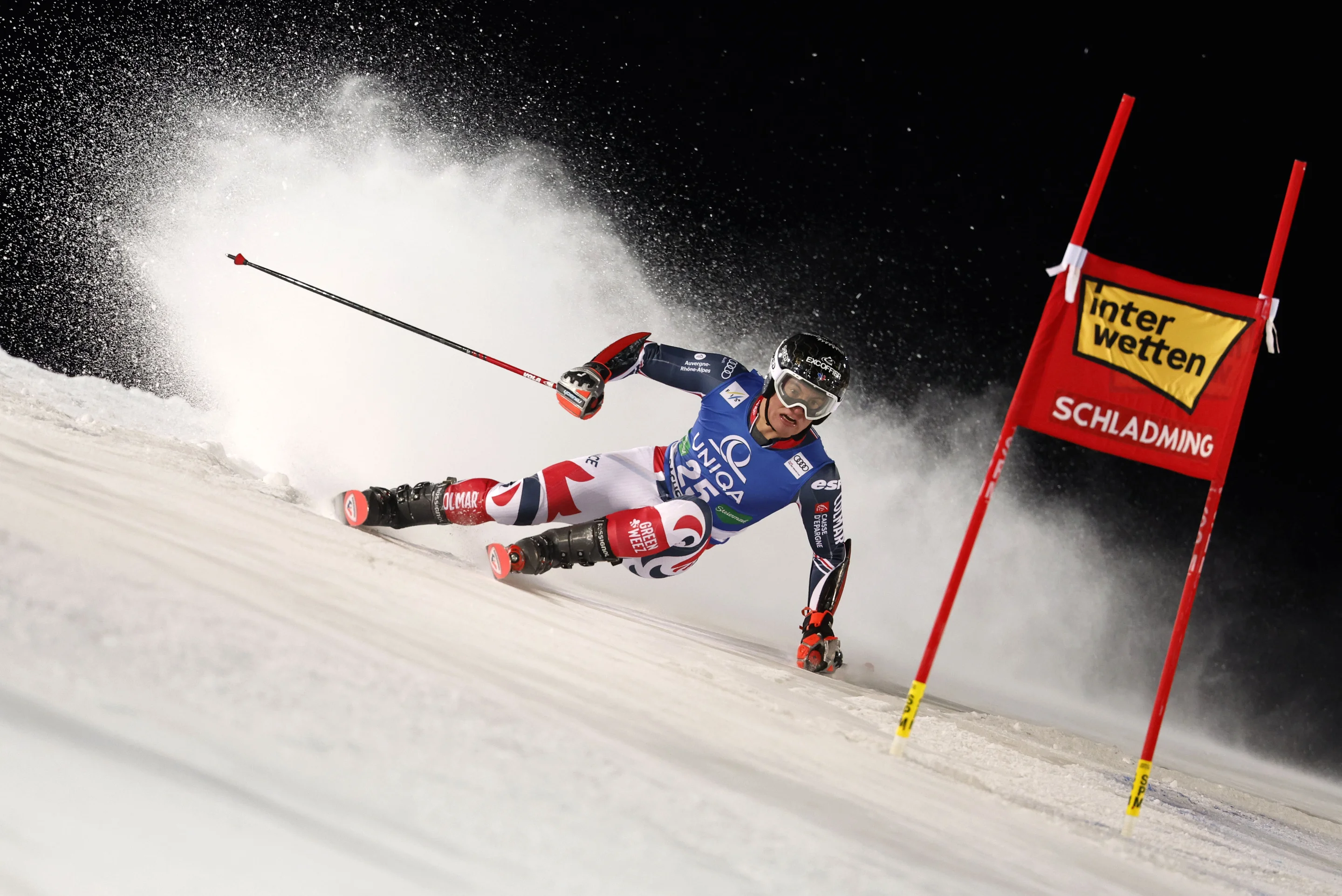A skier in a red and blue suit navigates a snowy slope at high speed, passing a red gate with "Schladming" under a night sky.