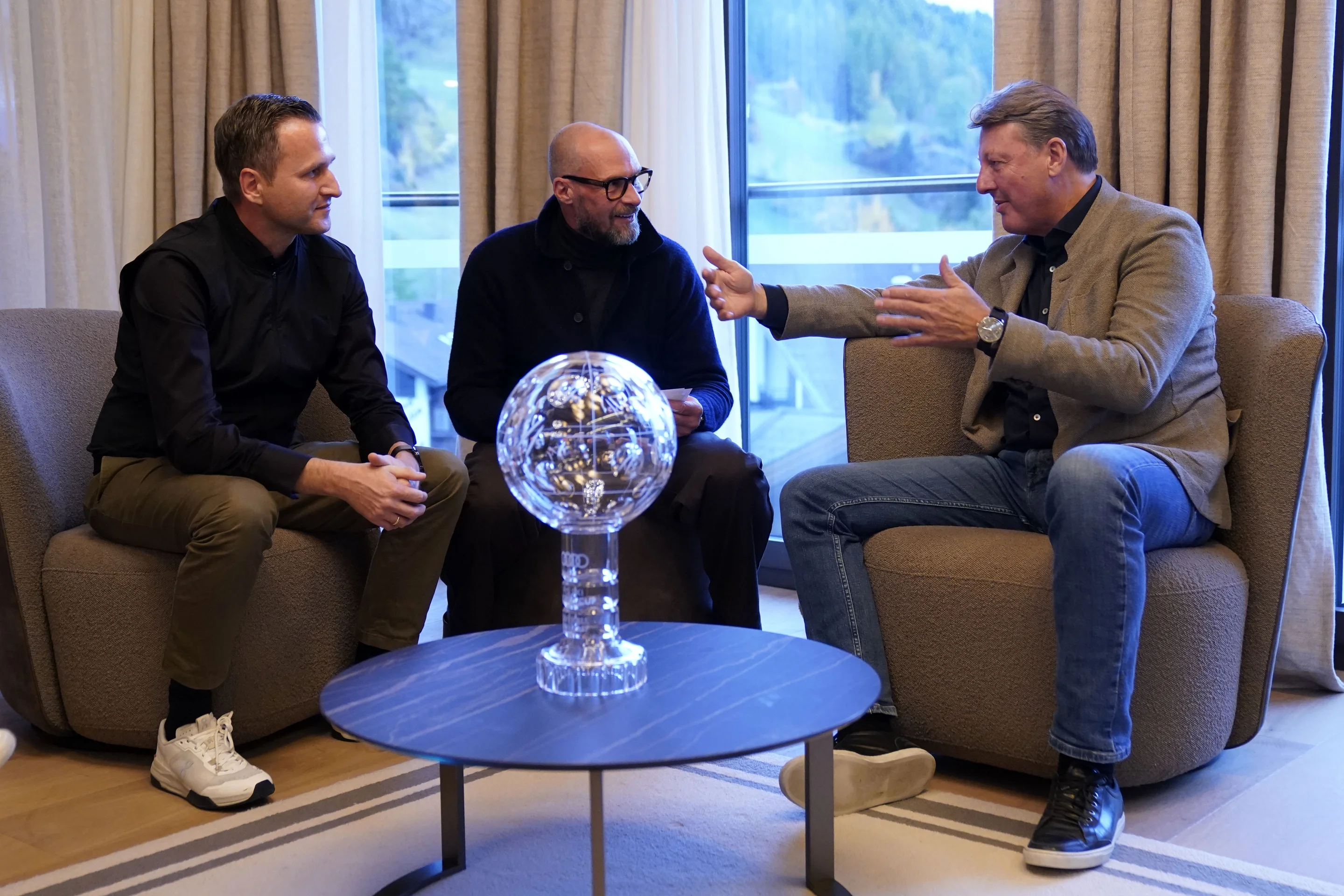 Three men sit around a small table with a glass globe centerpiece, engaged in animated conversation in a well-lit room.