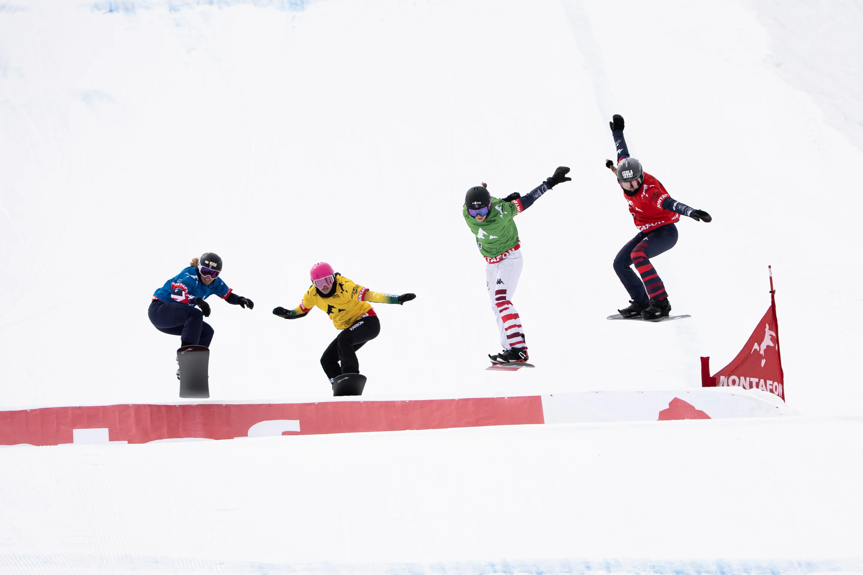 Four female snowboarders in blue, yellow, green and red bibs get some air over a jump with a big red ‘Montafon’ sign on it