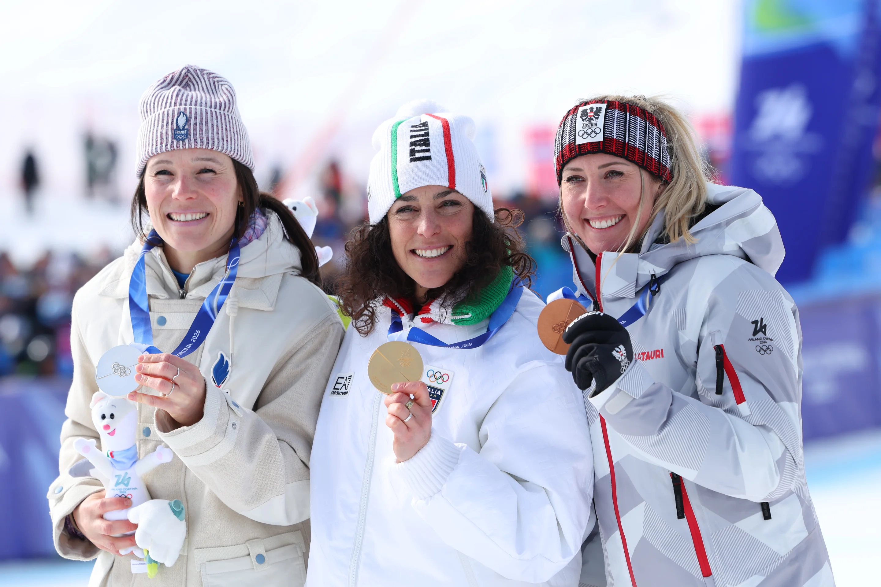 Three athletes at a winter event, each holding a medal and wearing winter gear, smile for the camera. Snowy background with event banners visible.