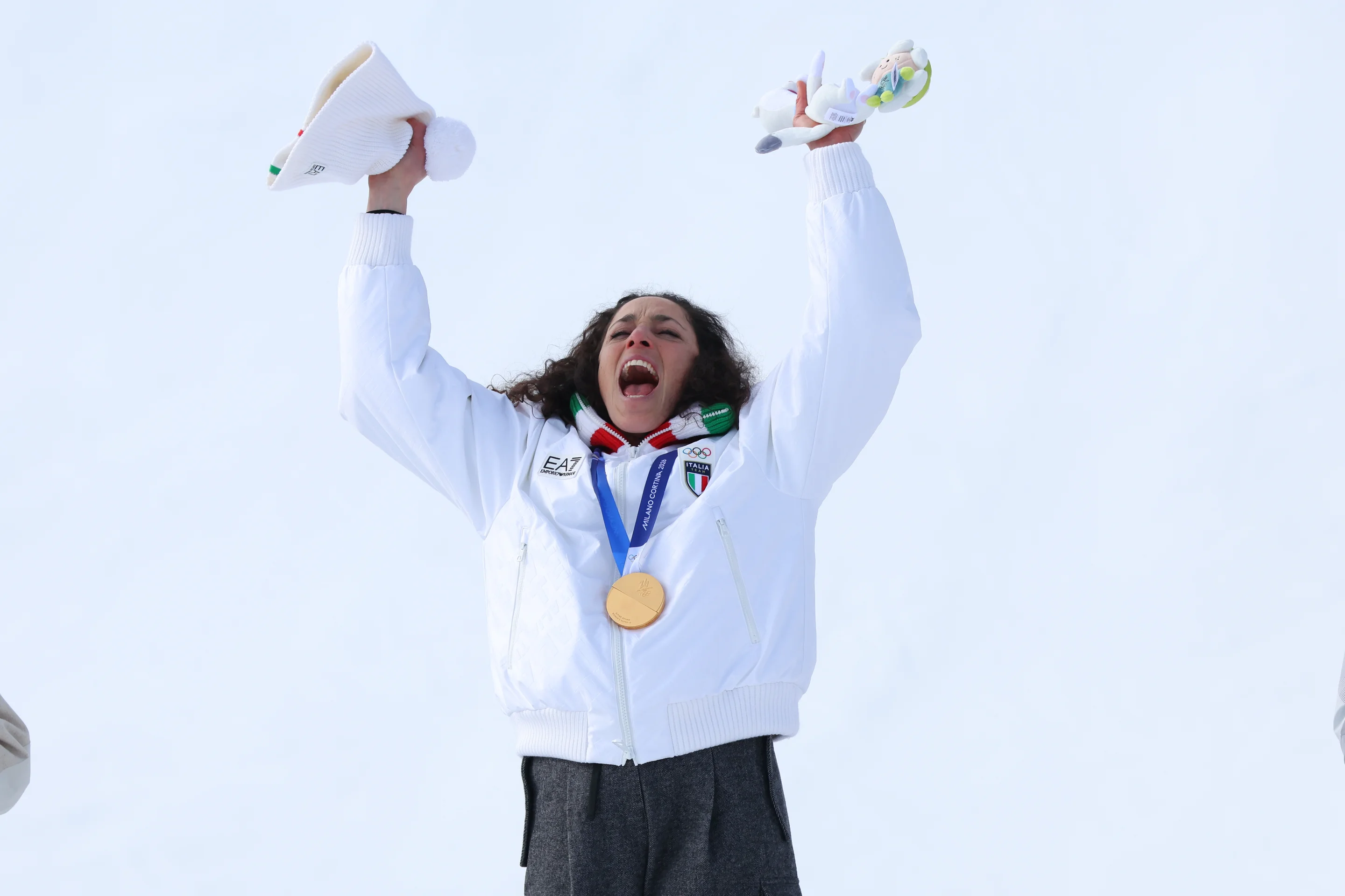 Athlete celebrating with arms raised, wearing a gold medal and holding a mascot, against a snowy background.