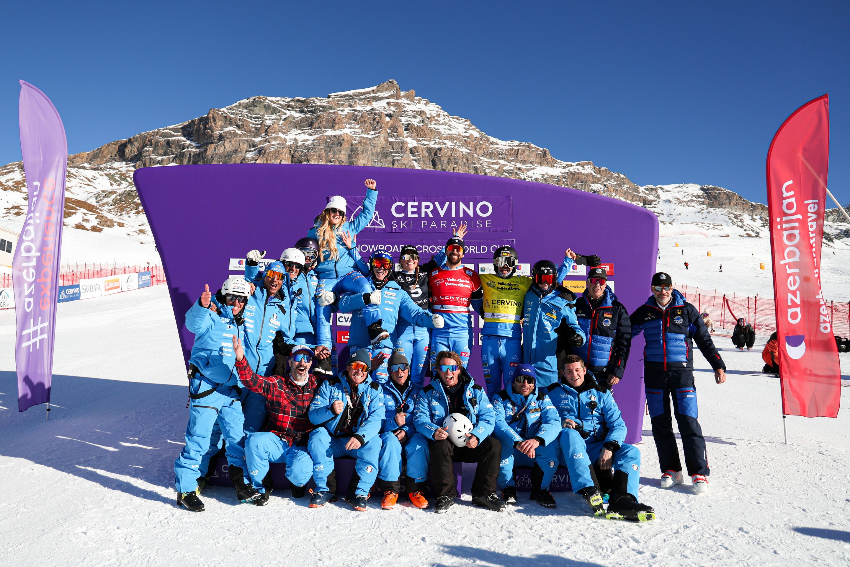 The Italian snowboard cross team, in blue snowsuits, lift Michela Moioli in the air in celebration. They stand in front of a purple backdrop labelled “Cervino Ski Paradise” with a rocky mountain in the background.