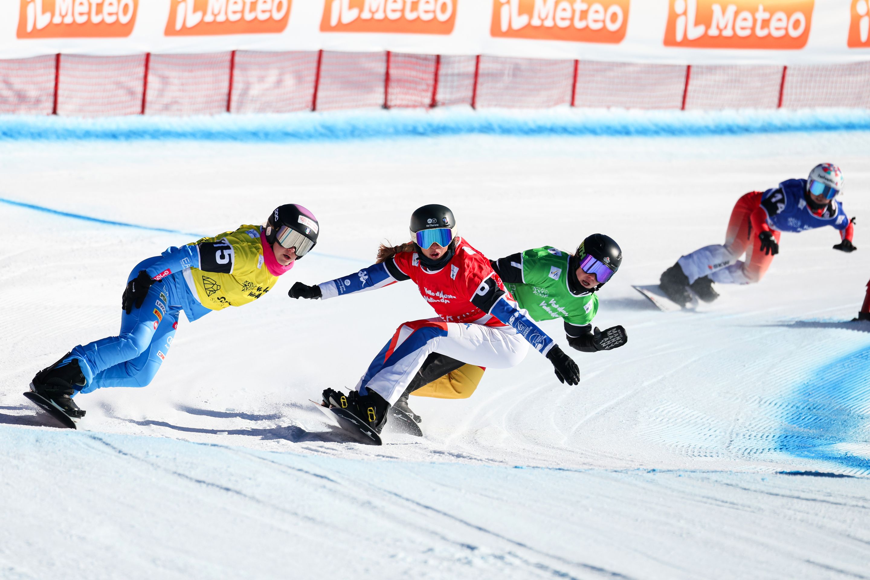 Four female snowboarders in yellow, red, green and blue bibs round a turn, with the rider in red, Chloe Trespeuch of France, leading. There is an orange crash barrier in the background.