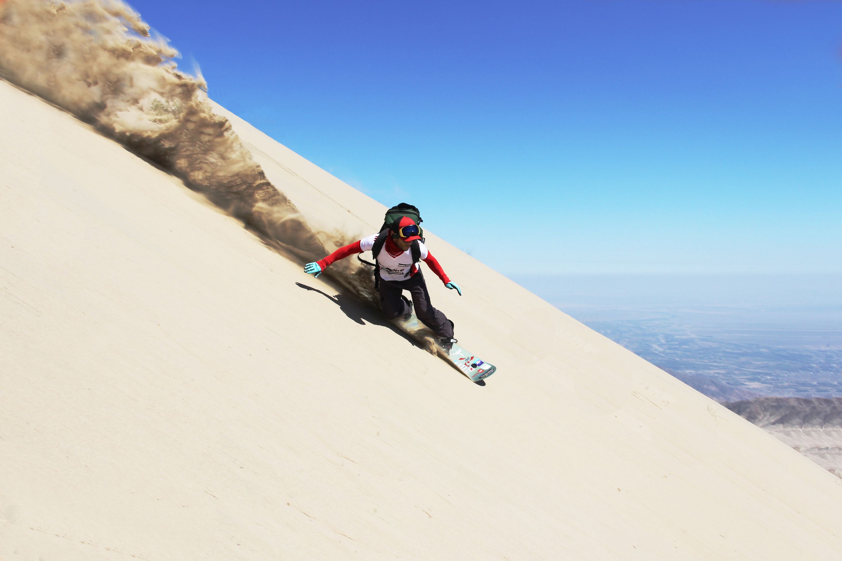 Dito Chavez sandboards down a dune at a steep angle, with sand flying out behind him and a view of blue sky and desert in the background