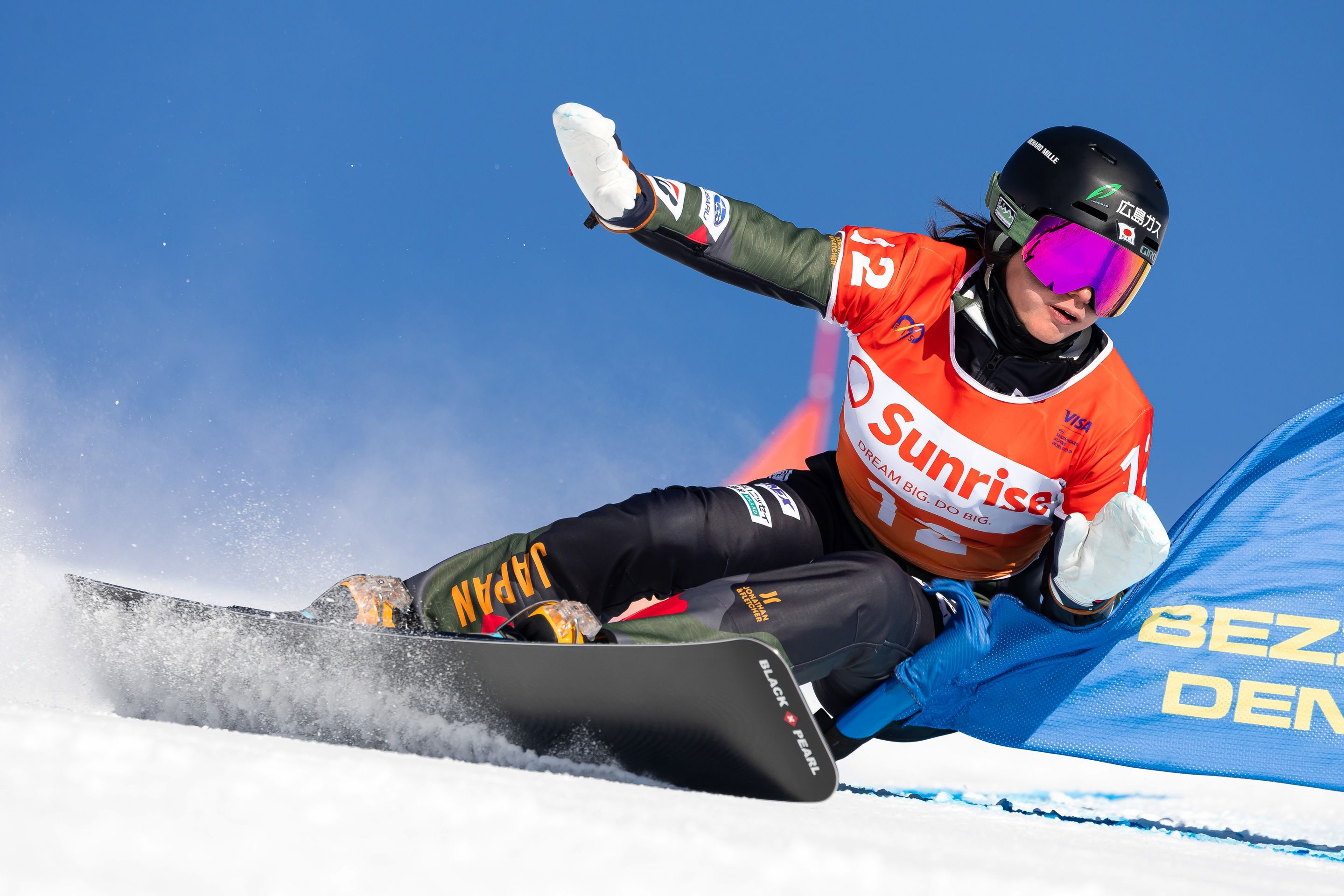 A snowboarder in an orange vest races downhill, kicking up snow, against a clear blue sky.