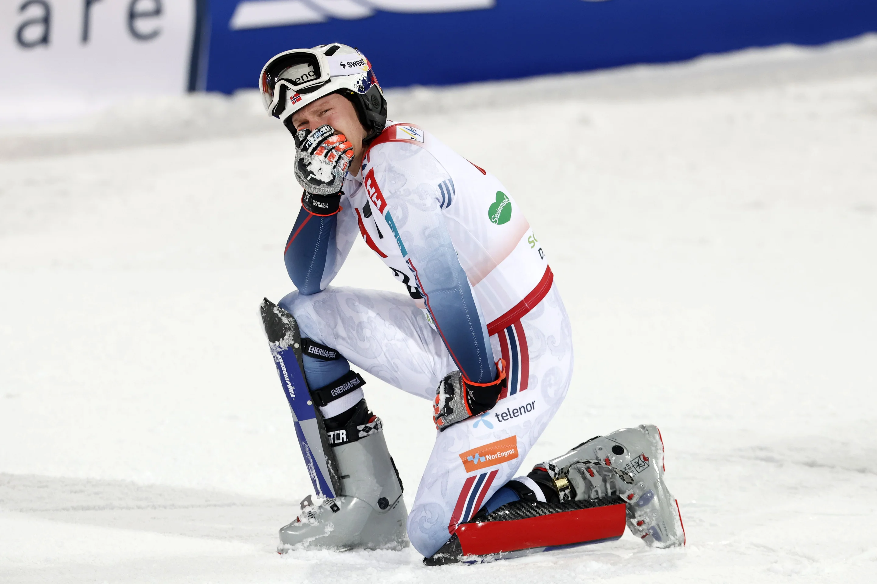 Skier kneeling on snow, wearing a white and red race suit, helmet, and goggles, with one hand on chin, appearing thoughtful or resting.