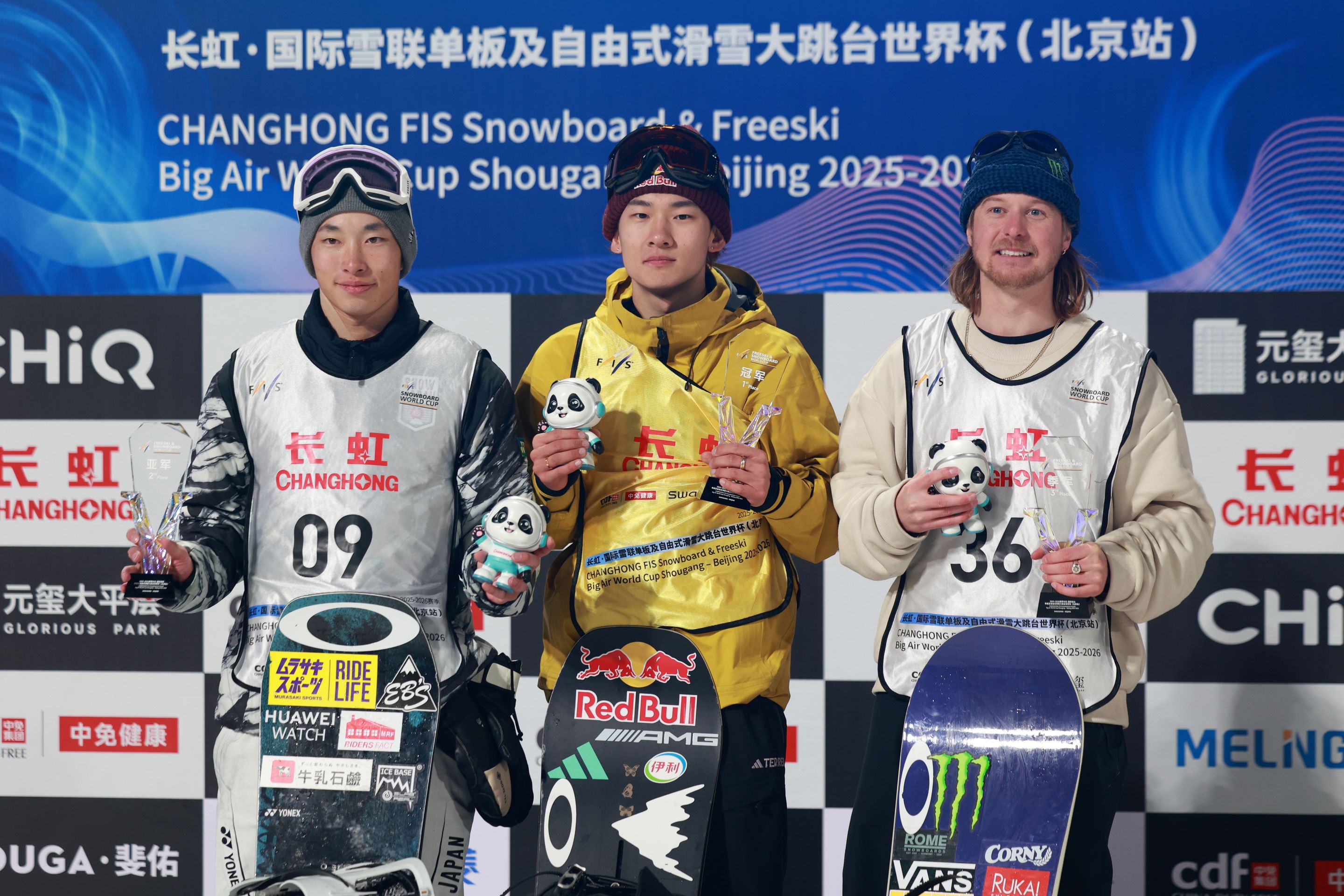 Three snowboarders on a podium holding panda mascots and snowboards; center winner in a yellow jacket under event signage.