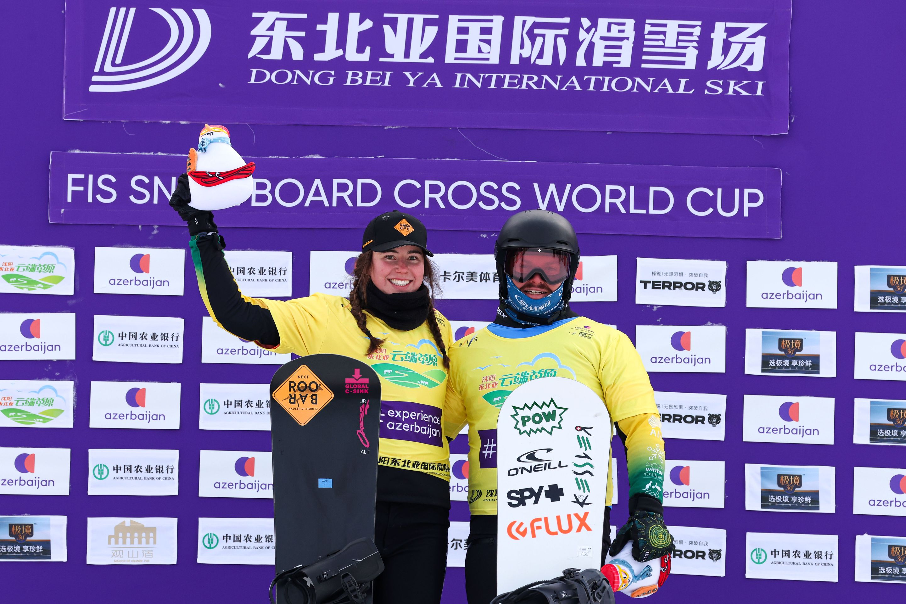 Two snowboarders in yellow jerseys pose with snowboards and helmets in front of a purple backdrop at a snowboarding event.