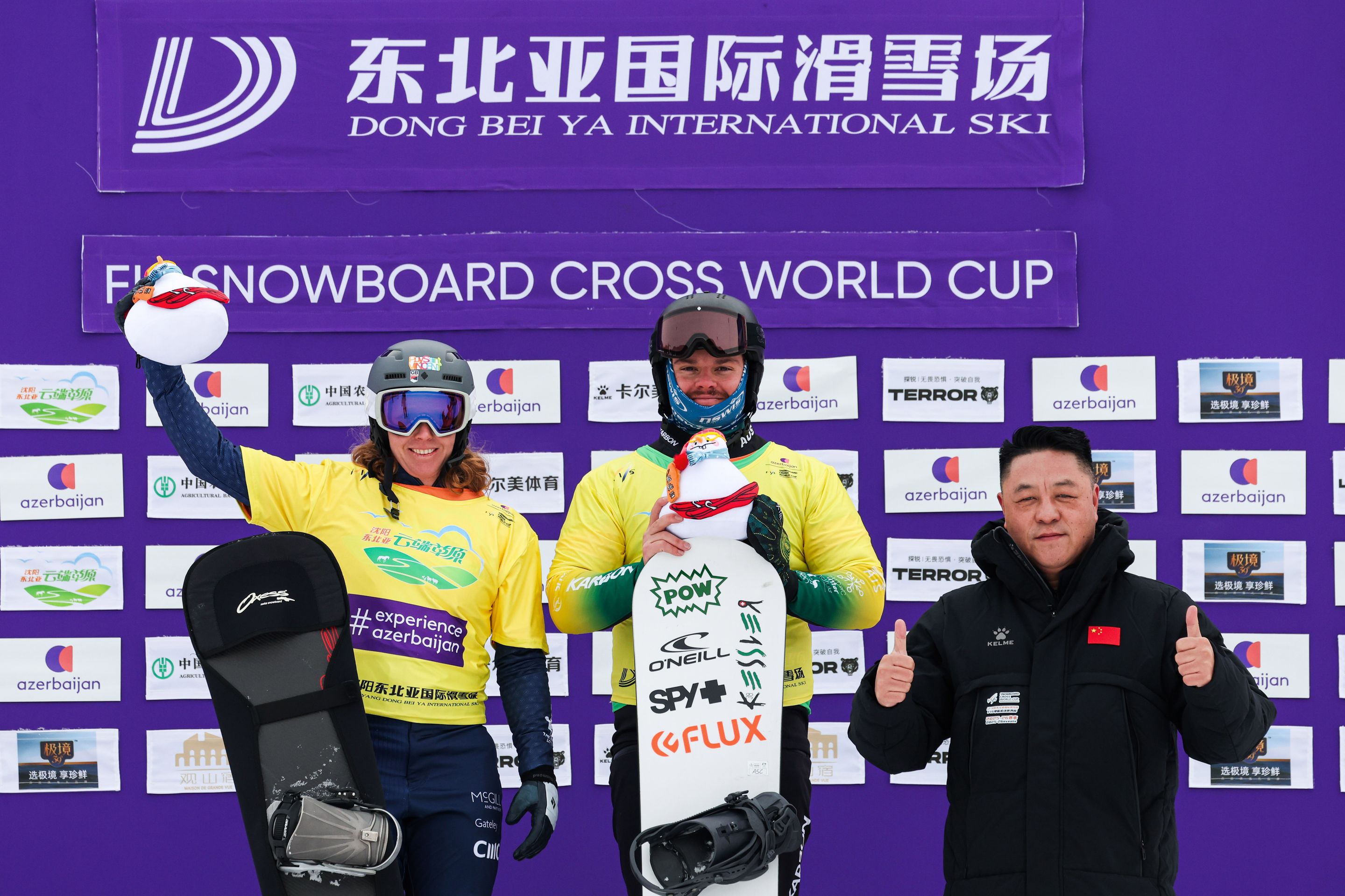 Snowboarders on podium celebrate at the FIS Snowboard Cross World Cup with an official, holding snowboards and wearing medals.