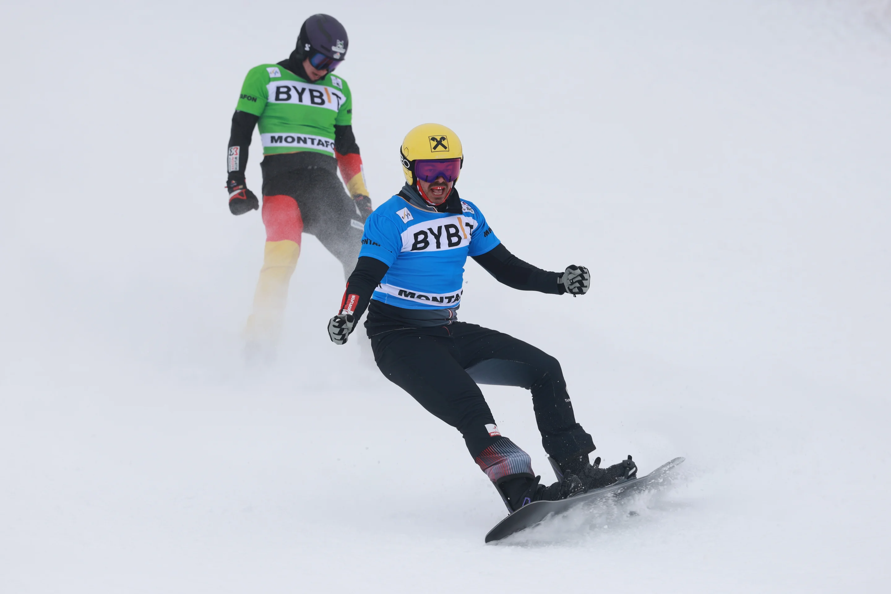 Snowboarder Jakob Dusek celebrates as he crosses the finish line. He is wearing a yellow helmet and a blue bib, with Leon Ulbricht in the background in a green bib.
