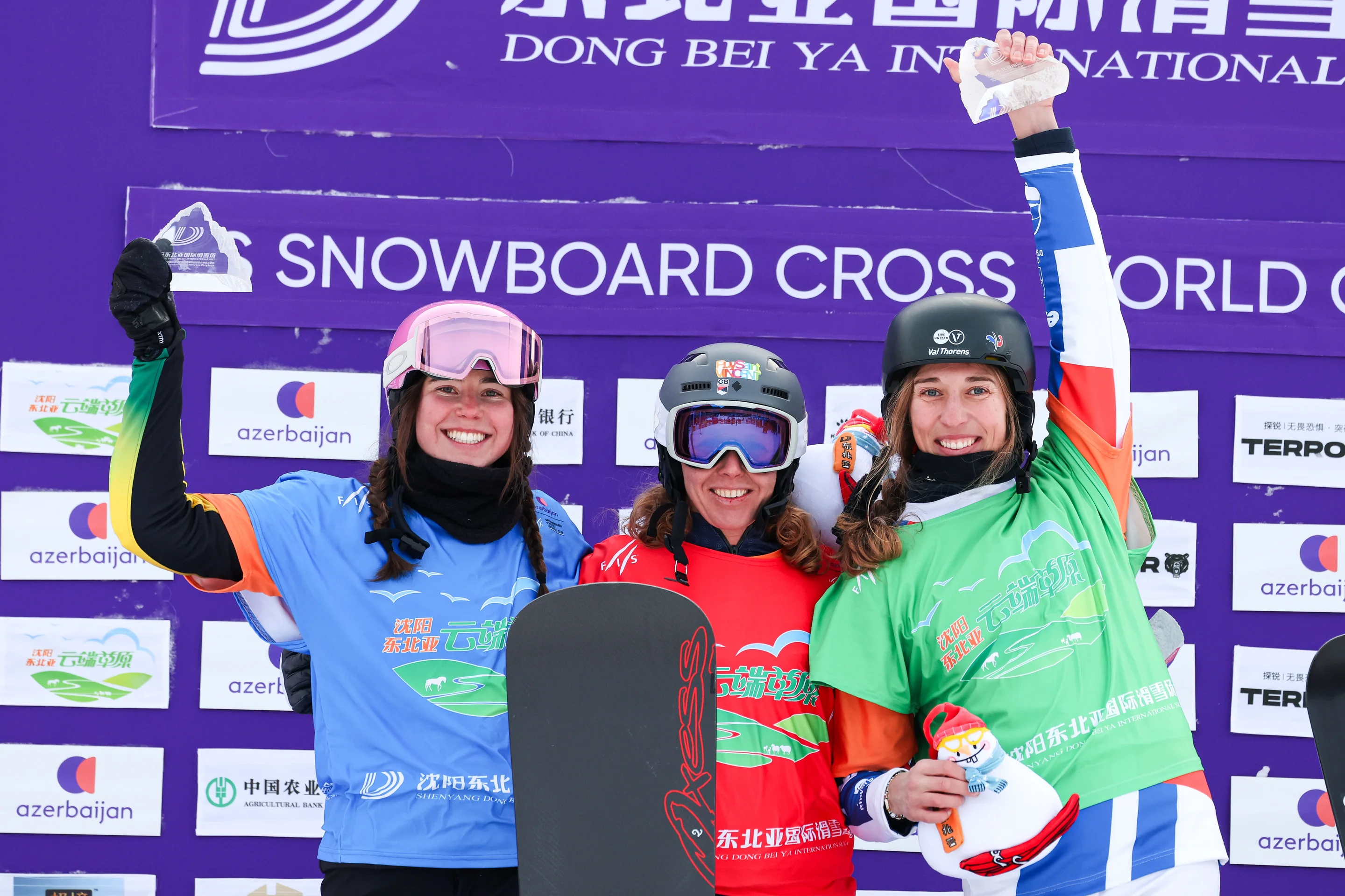 Three snowboarders posing on a podium, smiling widely, wearing colorful gear, with raised fists against a backdrop of a Snowboard Cross World Cup banner.