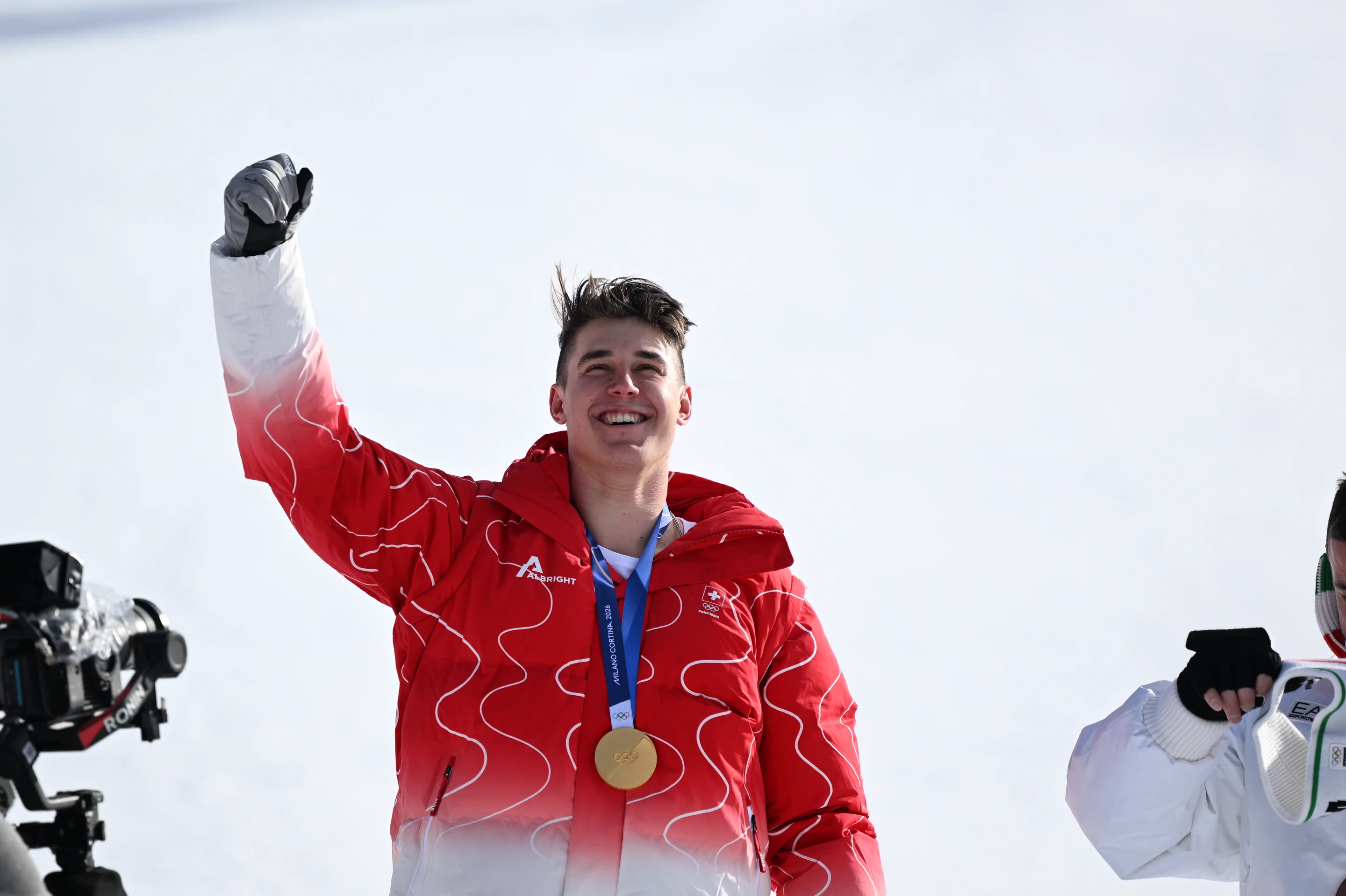 Athlete in a red jacket celebrates victory, holding a medal, with snow and a camera in the background.