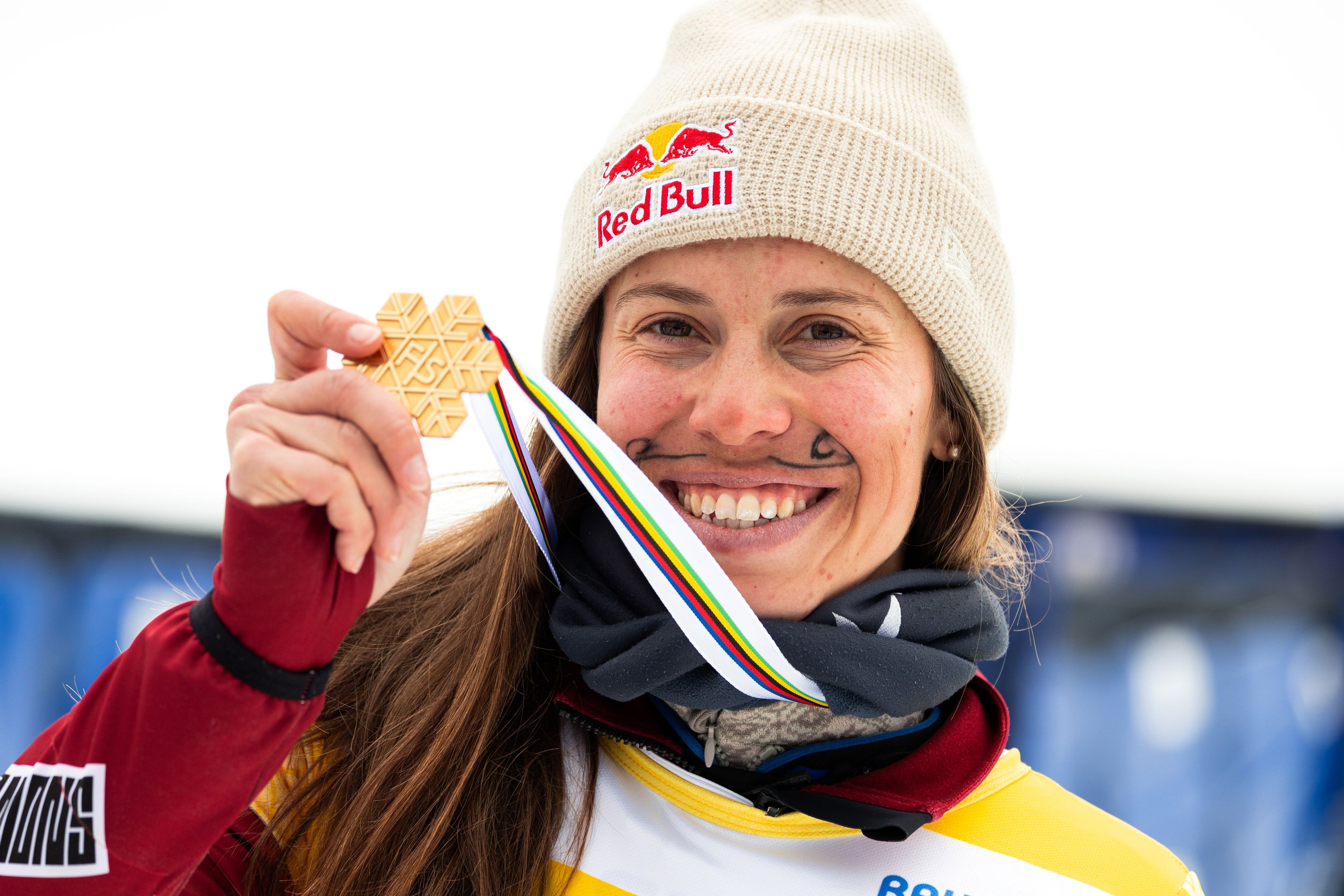 Eva Adamczykova, wearing a beige woolly hat with the Red Bull logo, holds up a gold medal shaped like a snowflake at the 2023 World Championships in Bakuriani