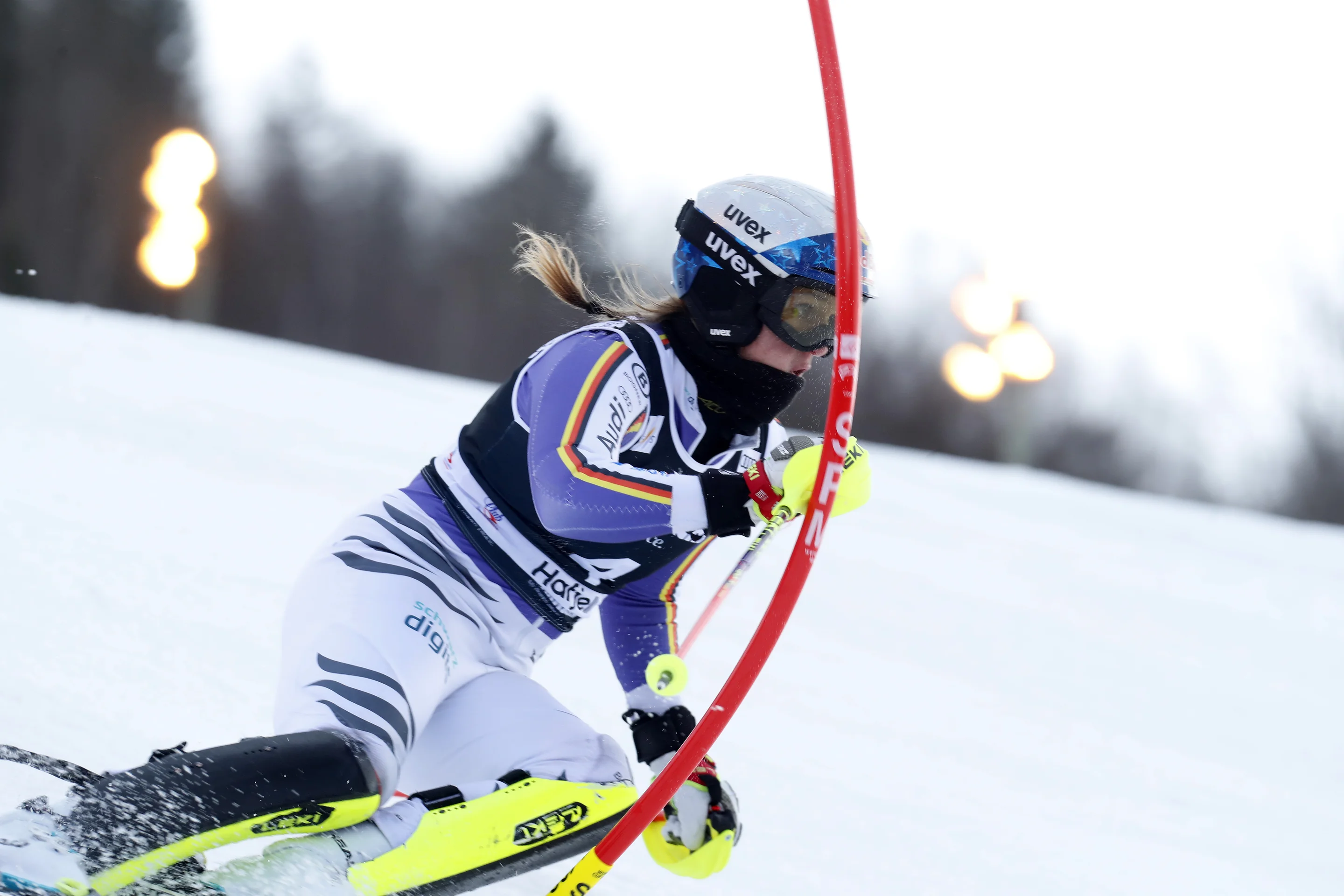 A skier in action on a snowy slope, wearing protective gear and a uniform, maneuvers past a red slalom pole under bright lights.