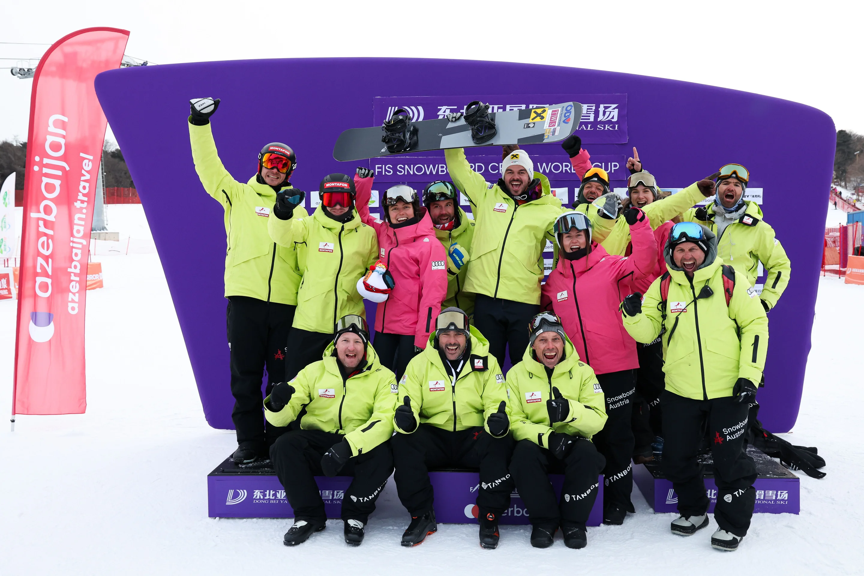 A group of snowboarders in bright jackets celebrate enthusiastically, posing in front of a purple podium with flags in the background.