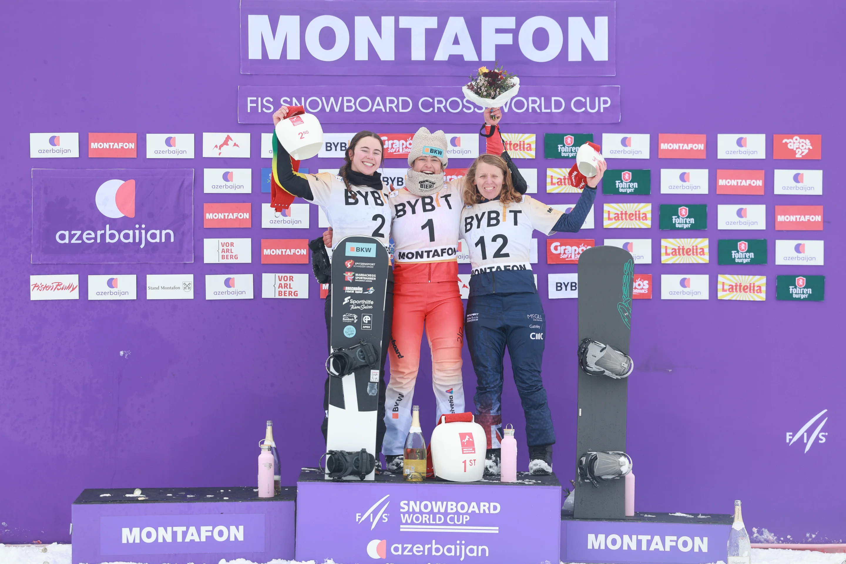 Josie Baff, Sina Siegenthaler and Charlotte Bankes stand on a purple podium in Montafon, lifting giant cowbells and a bunch of flowers in the air in celebration